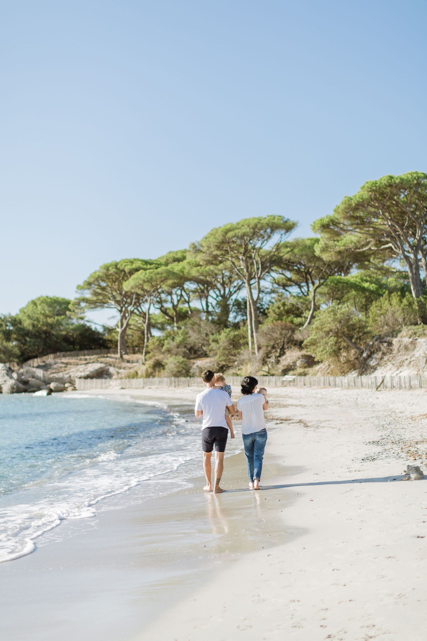 Une famille de trois personnes se promène dans la main le long d'une plage de sable blanc ensoleillée, avec des pins à Palombaggia, Corse.