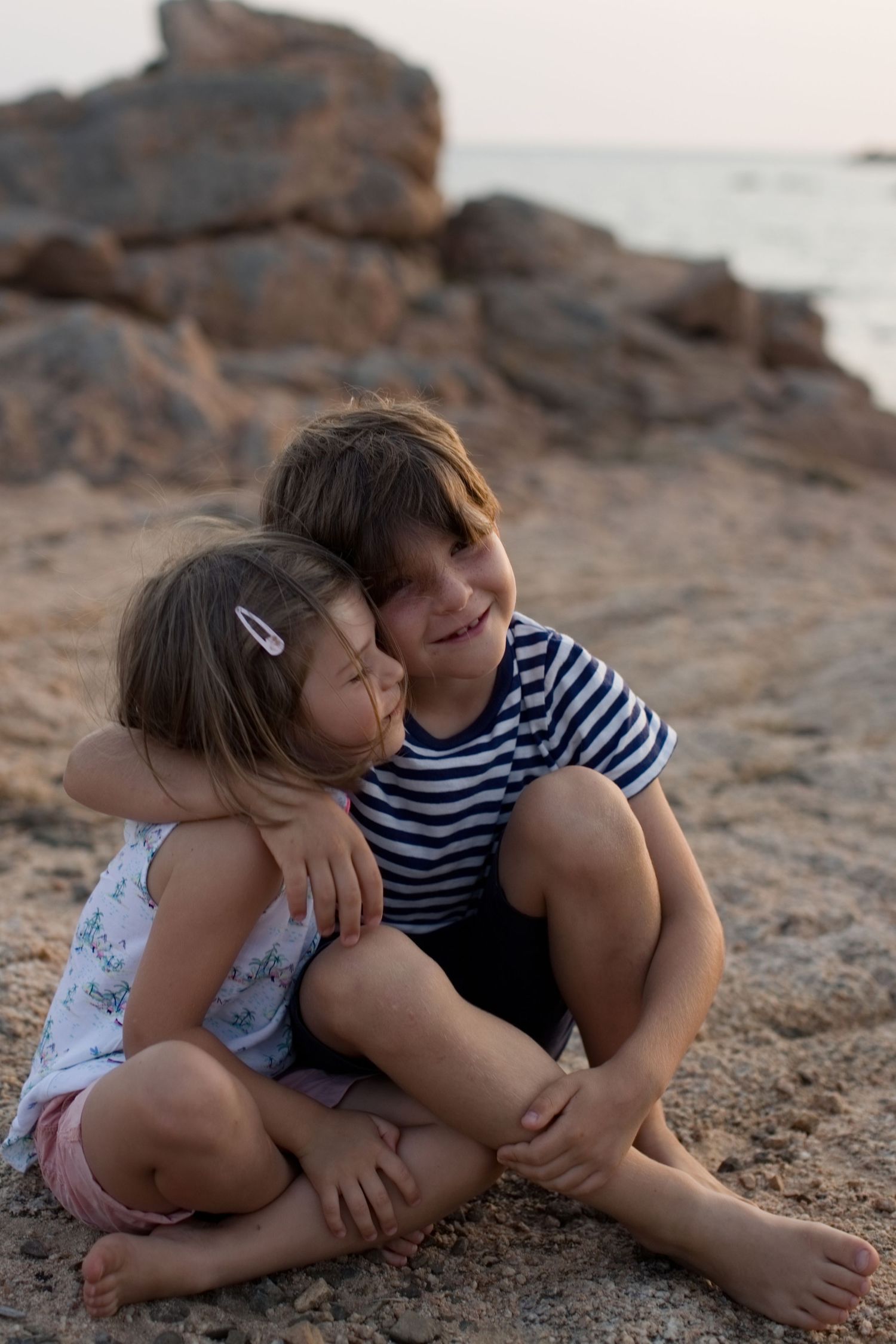 Deux jeunes frères et sœurs assis côte à côte sur une plage, s'enlaçant tendrement près des rivages rocheux à Bonifacio