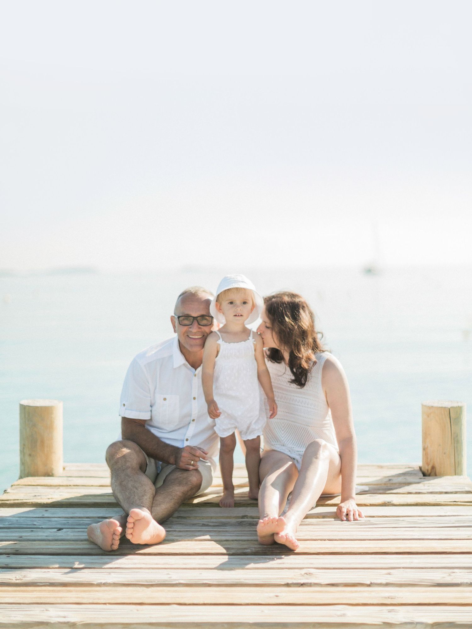 Une famille avec un bébé est assise sur une jetée ensoleillée, avec l'océan turquoise en arrière-plan. Sur la plage de Santa giulia.