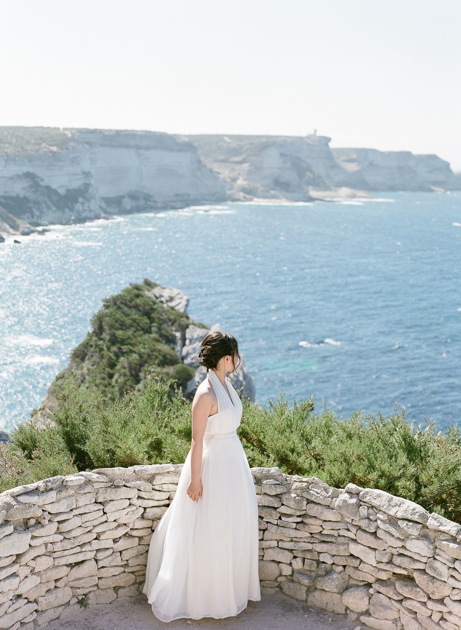 La mariée en robe blanche se tient sur un muret de pierre surplombant des falaises côtières spectaculaires et la mer Méditerranée