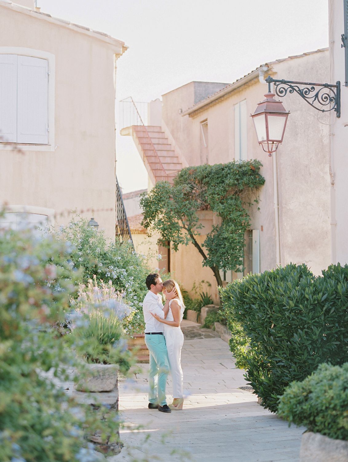 Un couple s'enlace dans une charmante ruelle d'un village français, entourée d'une végétation luxuriante et de chaleureux bâtiments