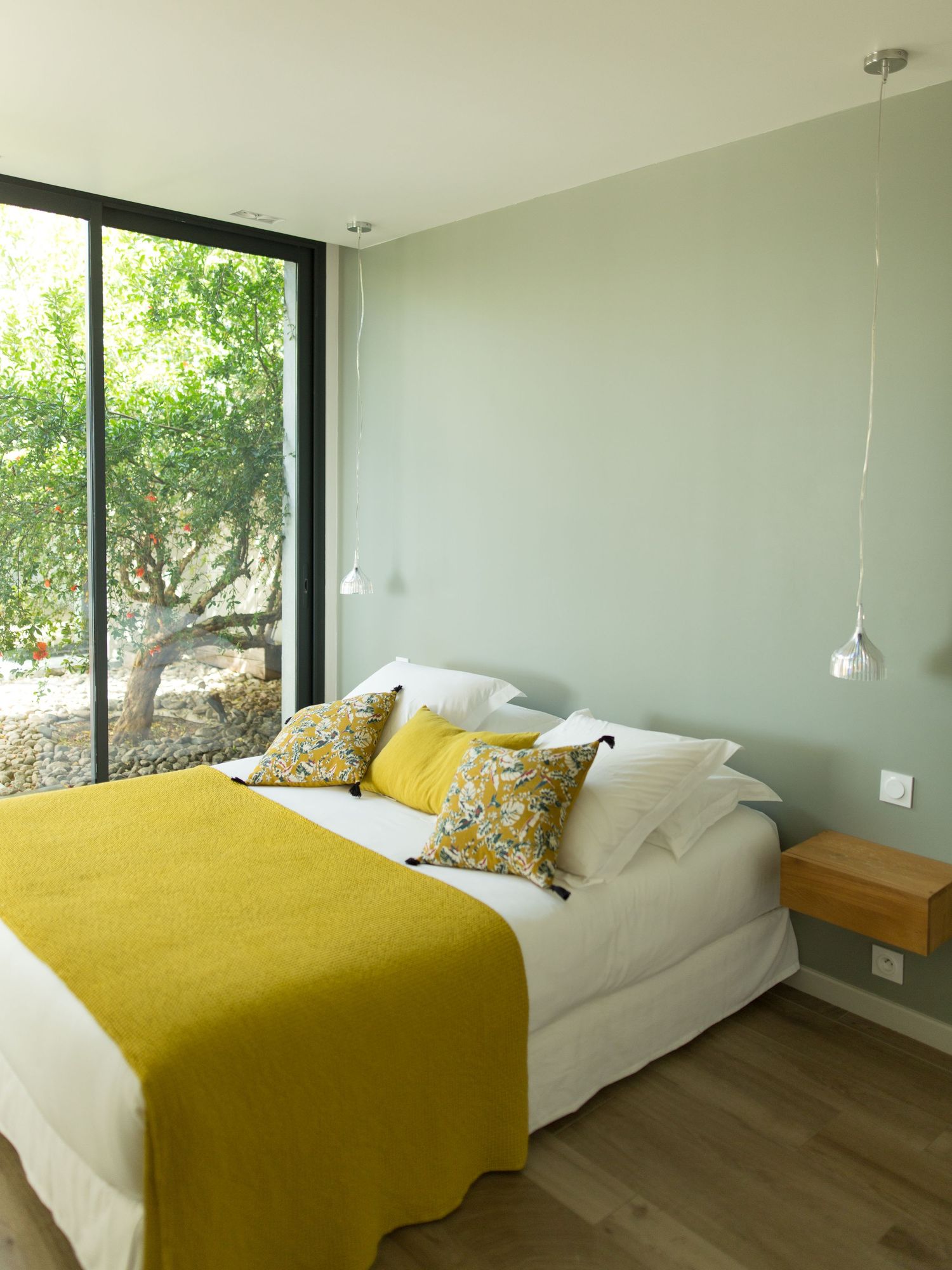 Chambre minimaliste avec murs vert sauge, couvre-lit jaune, coussins à motifs floraux, suspensions lumineuses.