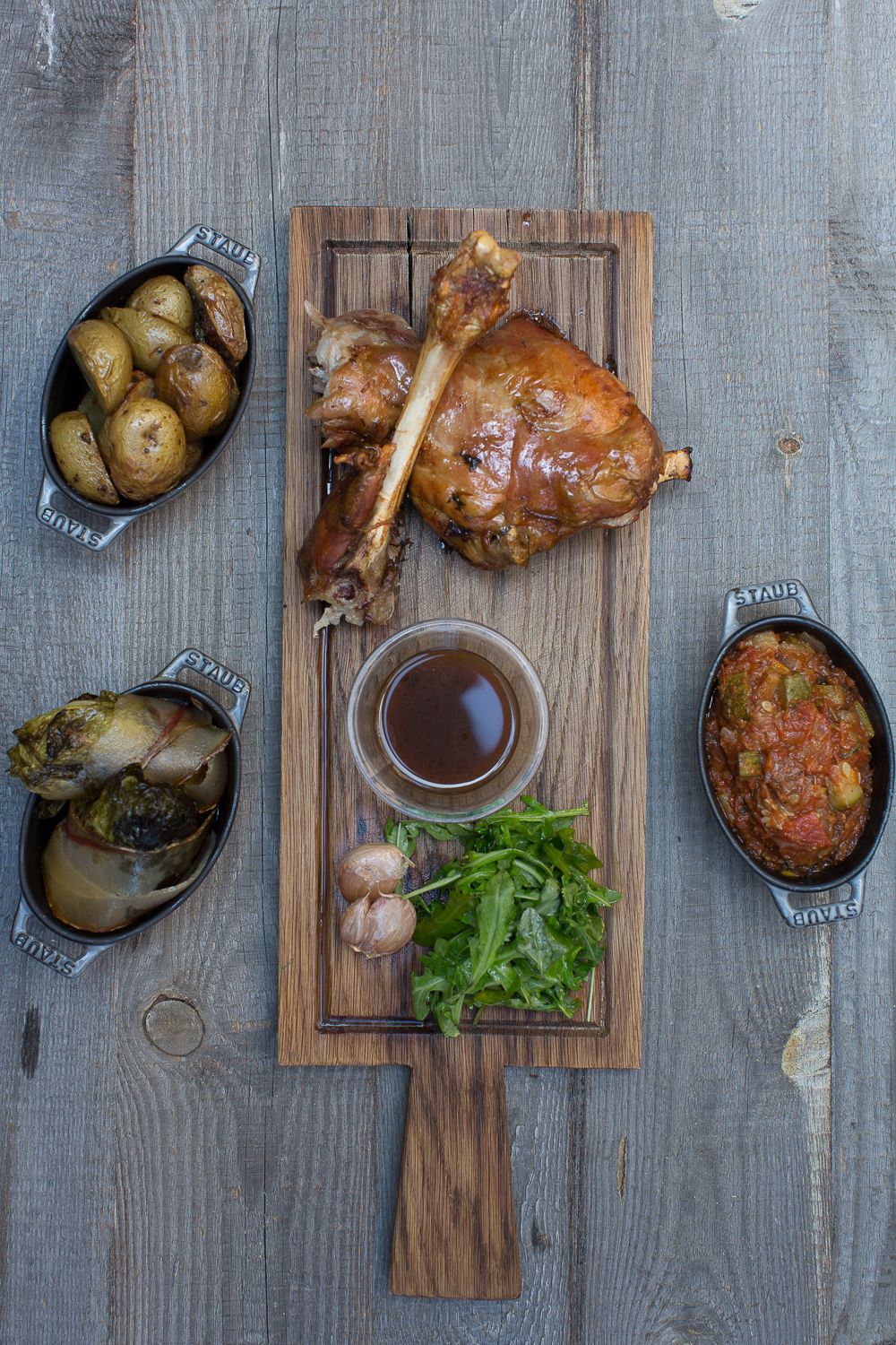 Jarret d'agneau rôti sur planche en bois, accompagné de pommes de terre, d'artichauts, de sauce tomate et d'herbes fraîches.