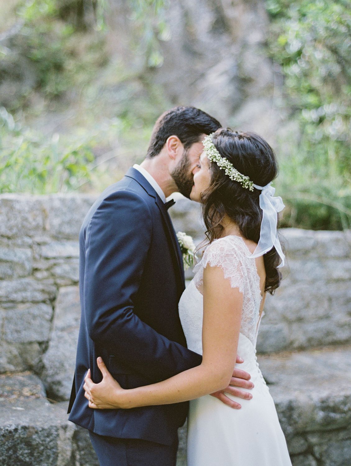 Bride and groom share a romantic forehead kiss outdoors, bride wearing lace dress and floral crown.
