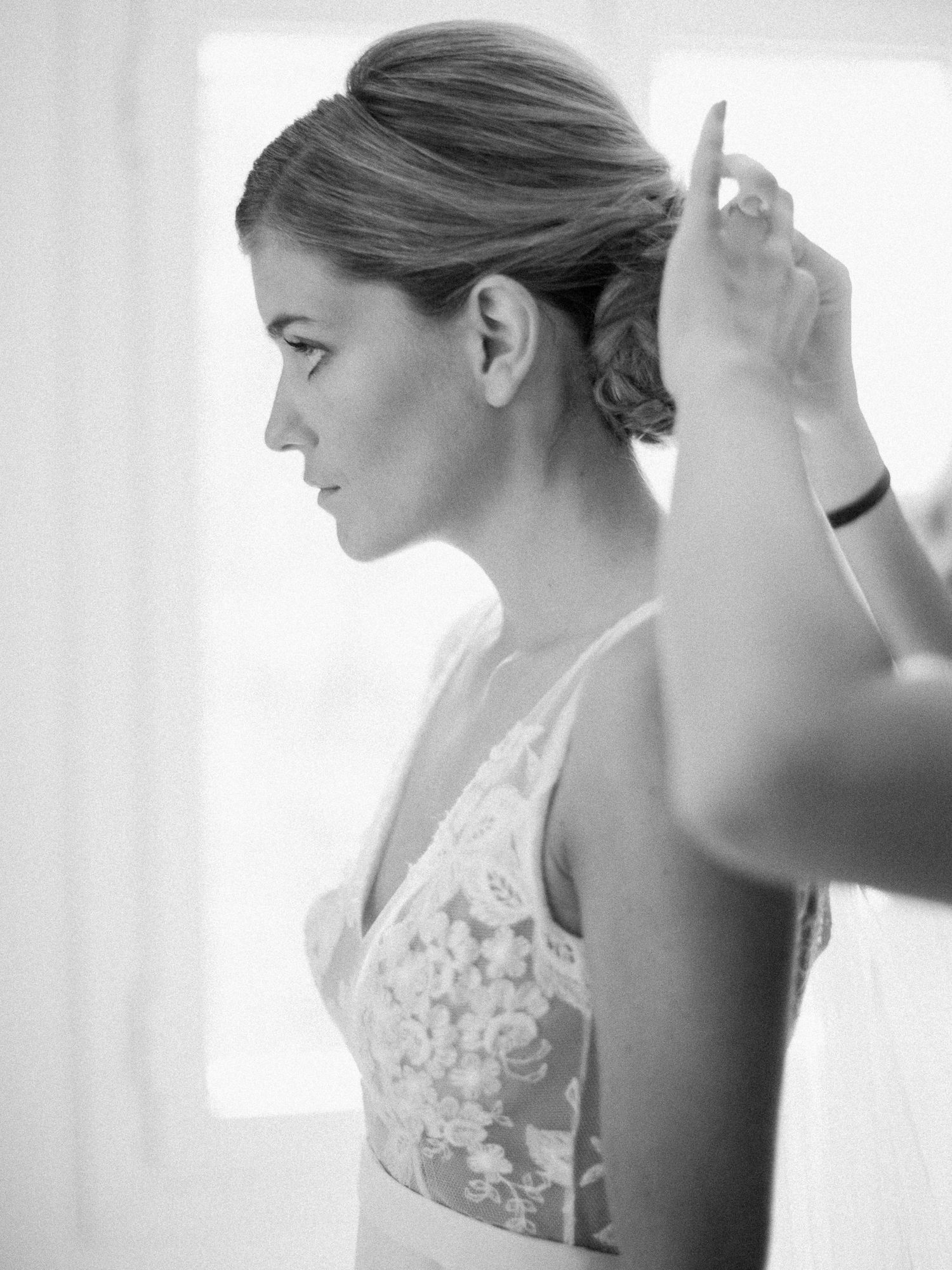 Une mariée se fait coiffer en un élégant chignon et porte une robe de mariée en dentelle florale ; photo en noir et blanc.