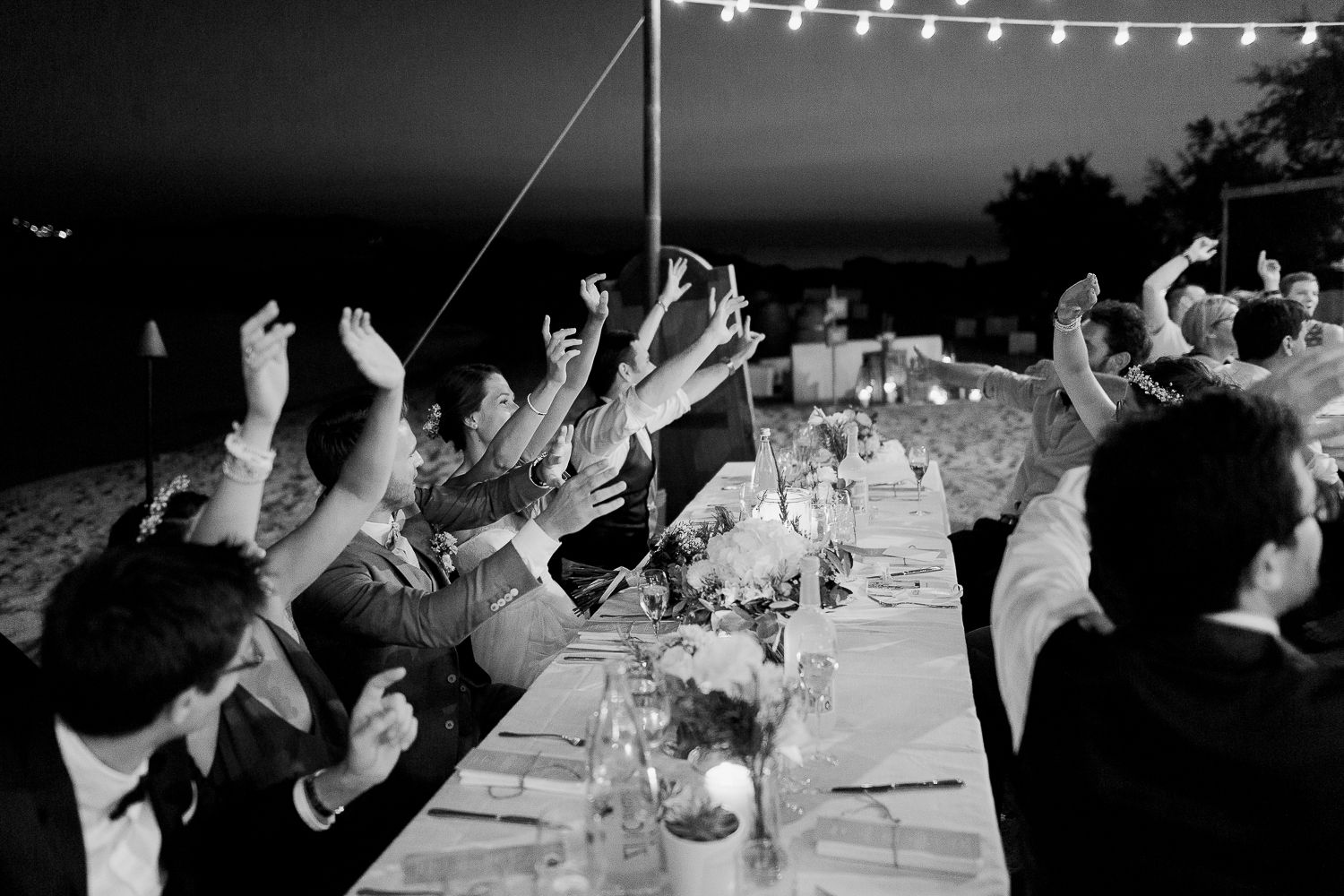 Dîner de réception de mariage en noir et blanc, les invités levant les mains sous le ciel étoilé, mariage à calvi paillote pain de sucre.