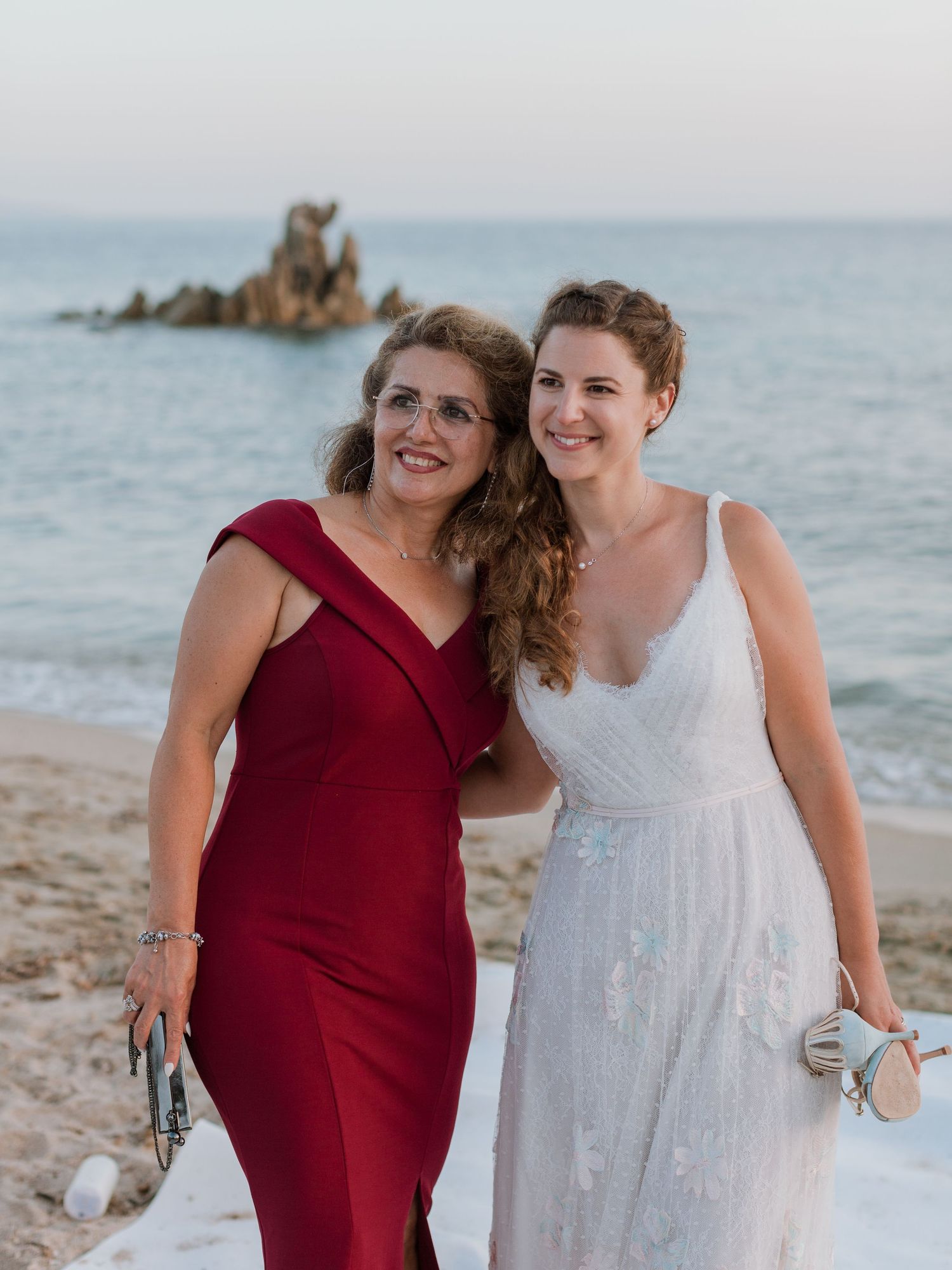 Deux femmes sourient sur une plage — l'une vêtue d'une robe rouge, l'autre d'une robe de mariée blanche — avec un paysage marin rocheux