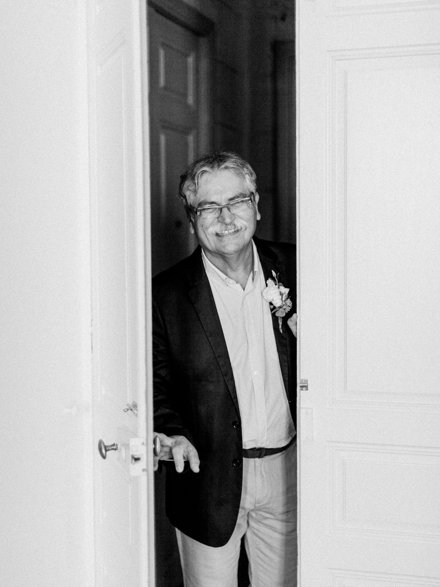 Man in blazer with boutonniere smiling through doorway in black and white wedding photo.