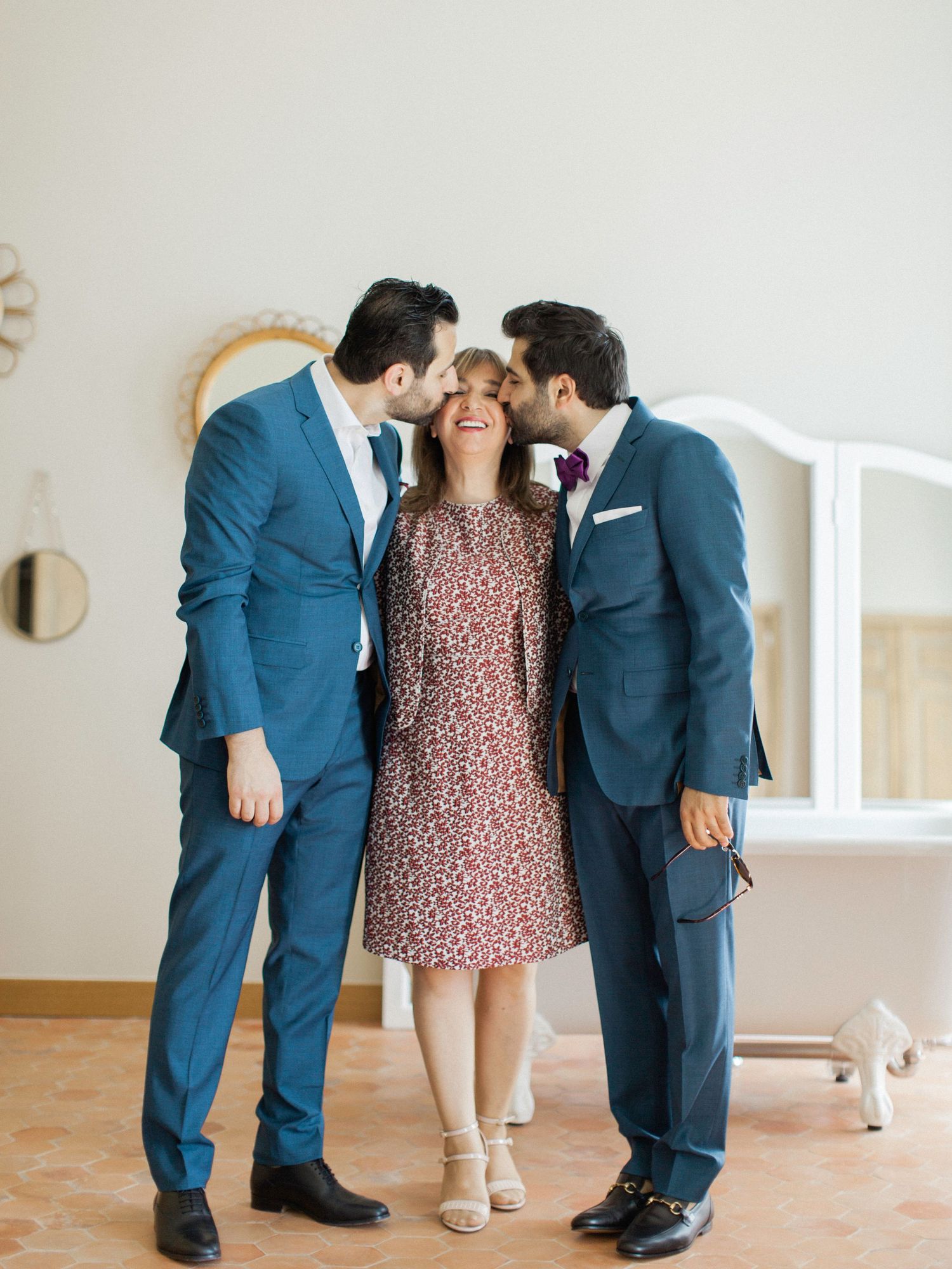 Two men in blue suits kissing a smiling woman in a floral dress on the cheeks indoors.