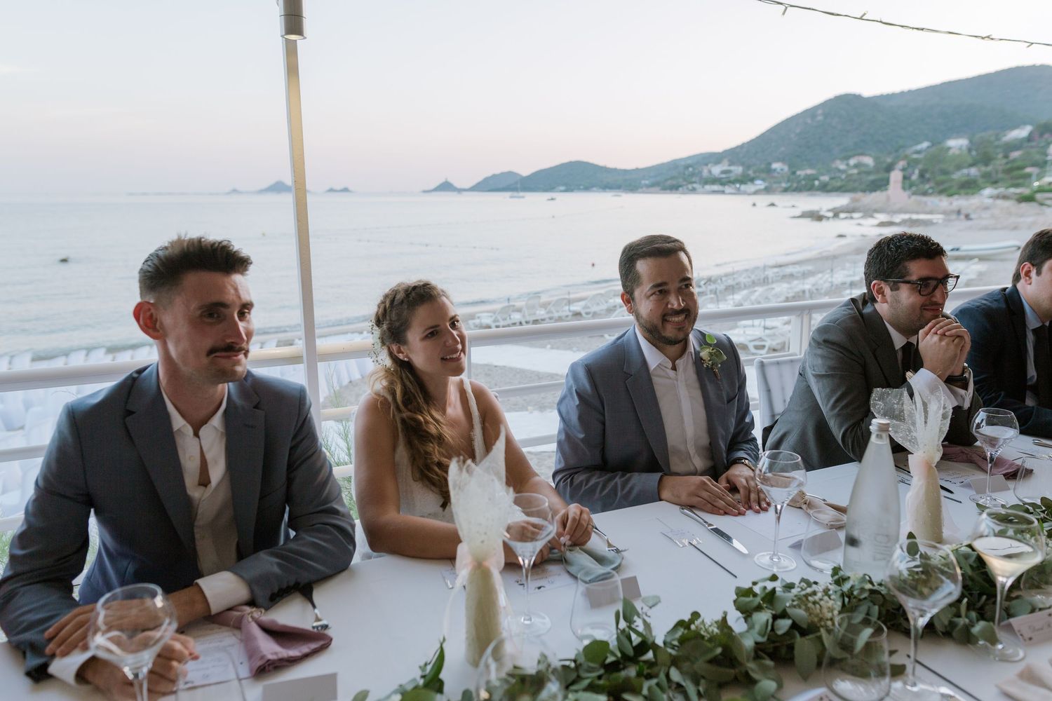Le cortège nuptial est assis à une table de réception en plein air, en bord de plage, avec vue sur l'océan au crépuscule.