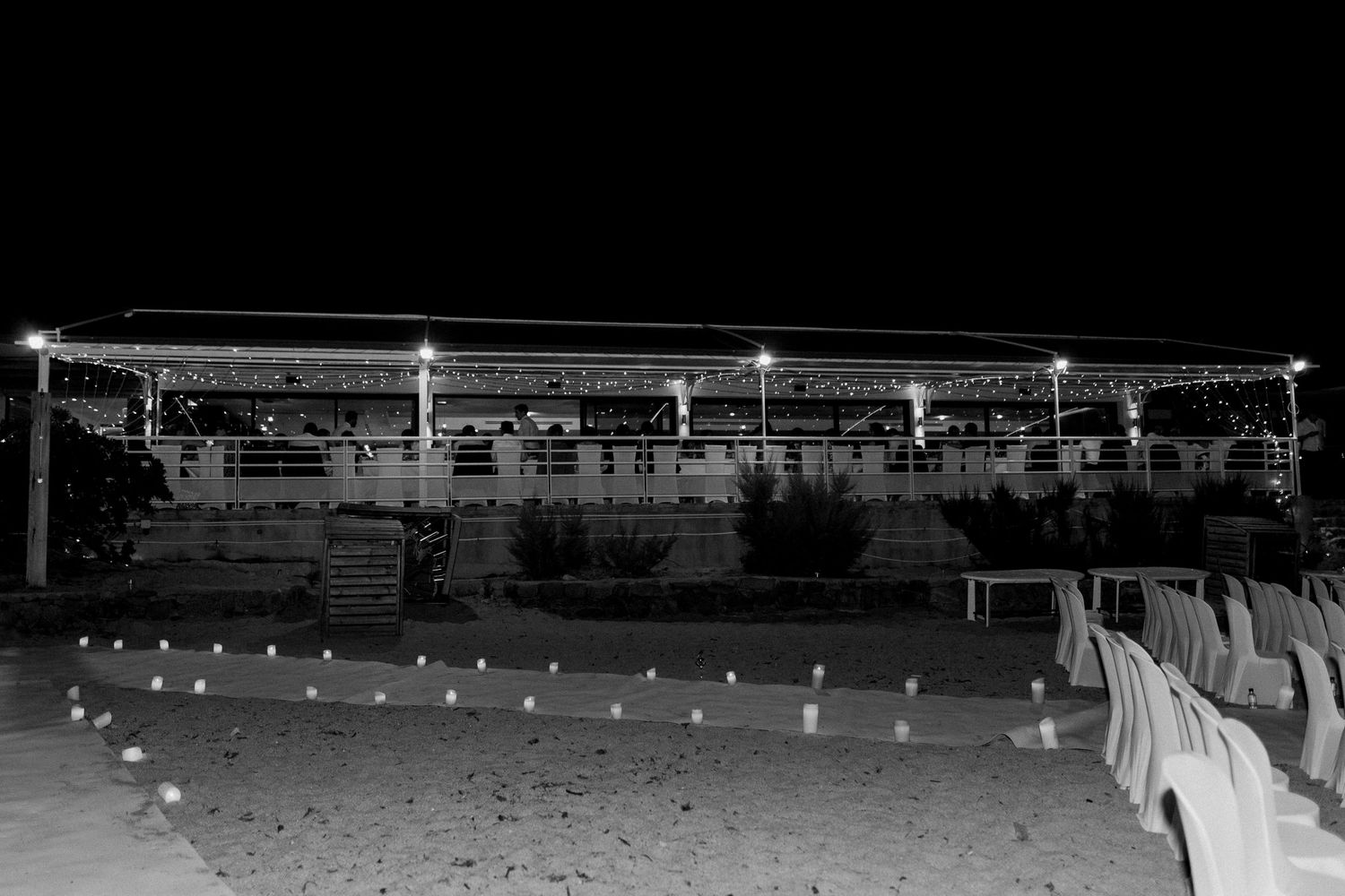 Photo nocturne en noir et blanc d'un stade extérieur illuminé, avec des bougies bordant des allées de sable.