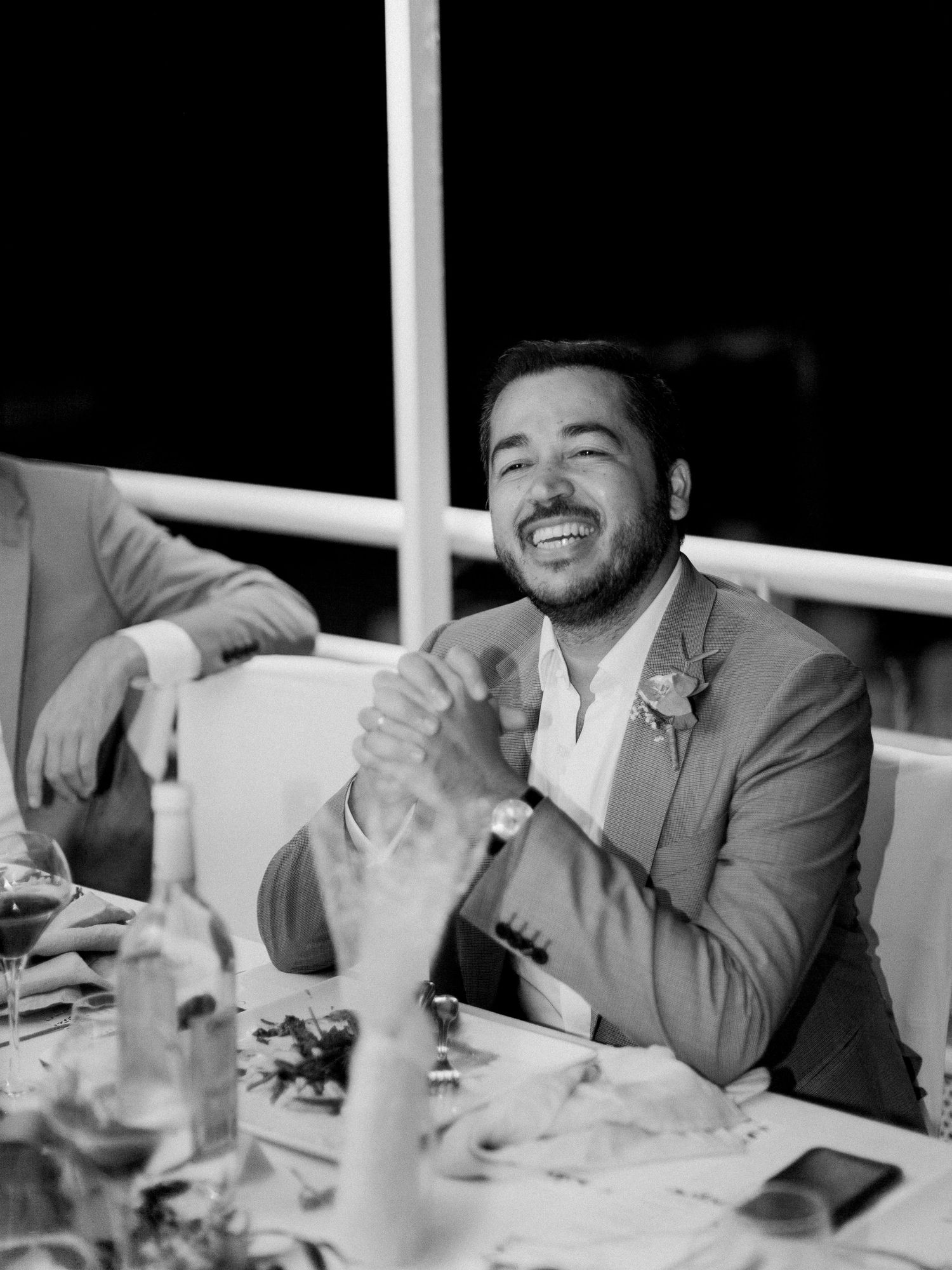 Un homme en costume gris rit joyeusement à une table de réception de mariage, sur une photographie en noir et blanc.