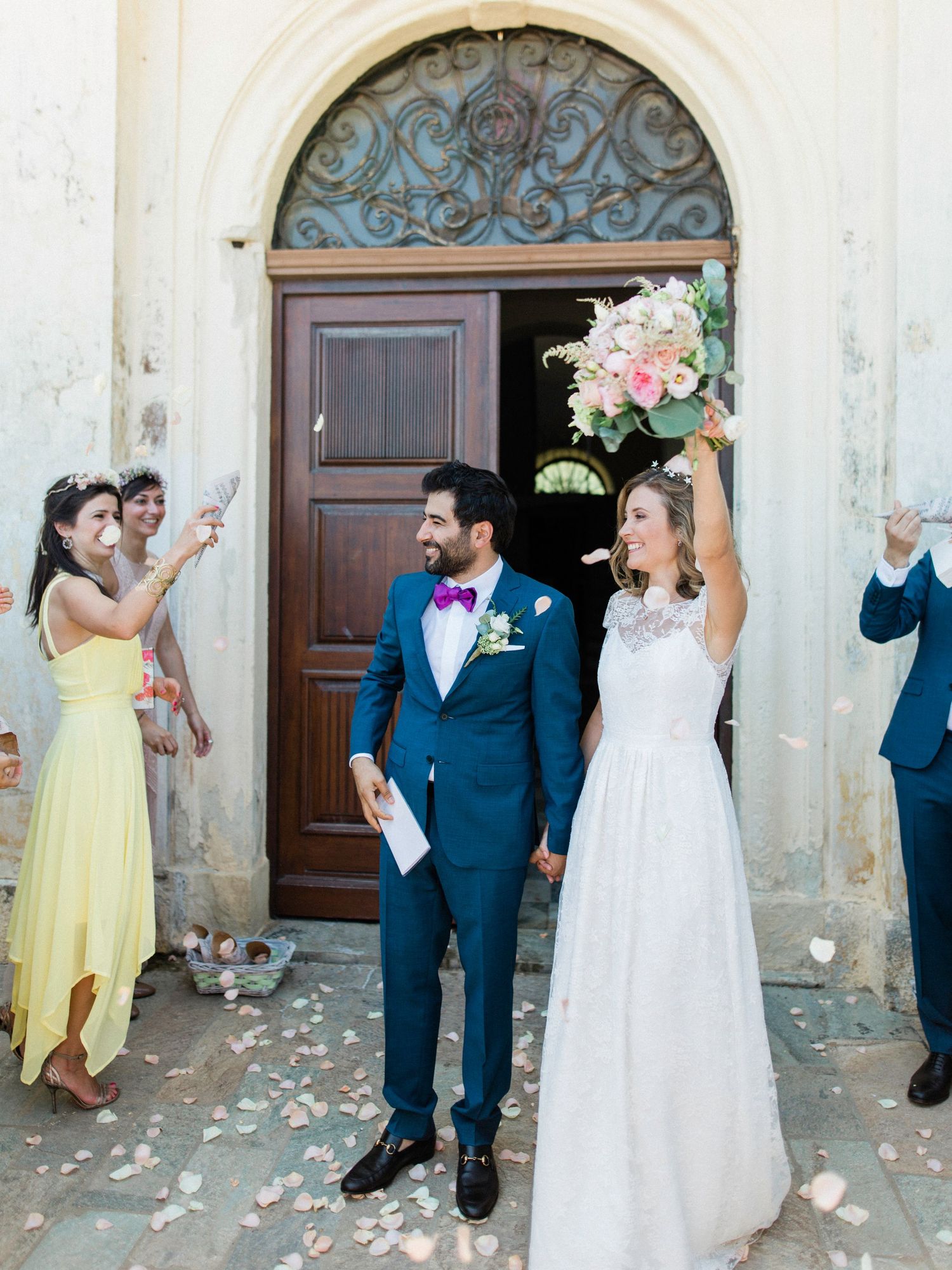 Newlywed couple celebrating outside a church, bride raising bouquet, groom in blue suit, guests throwing rose petals.