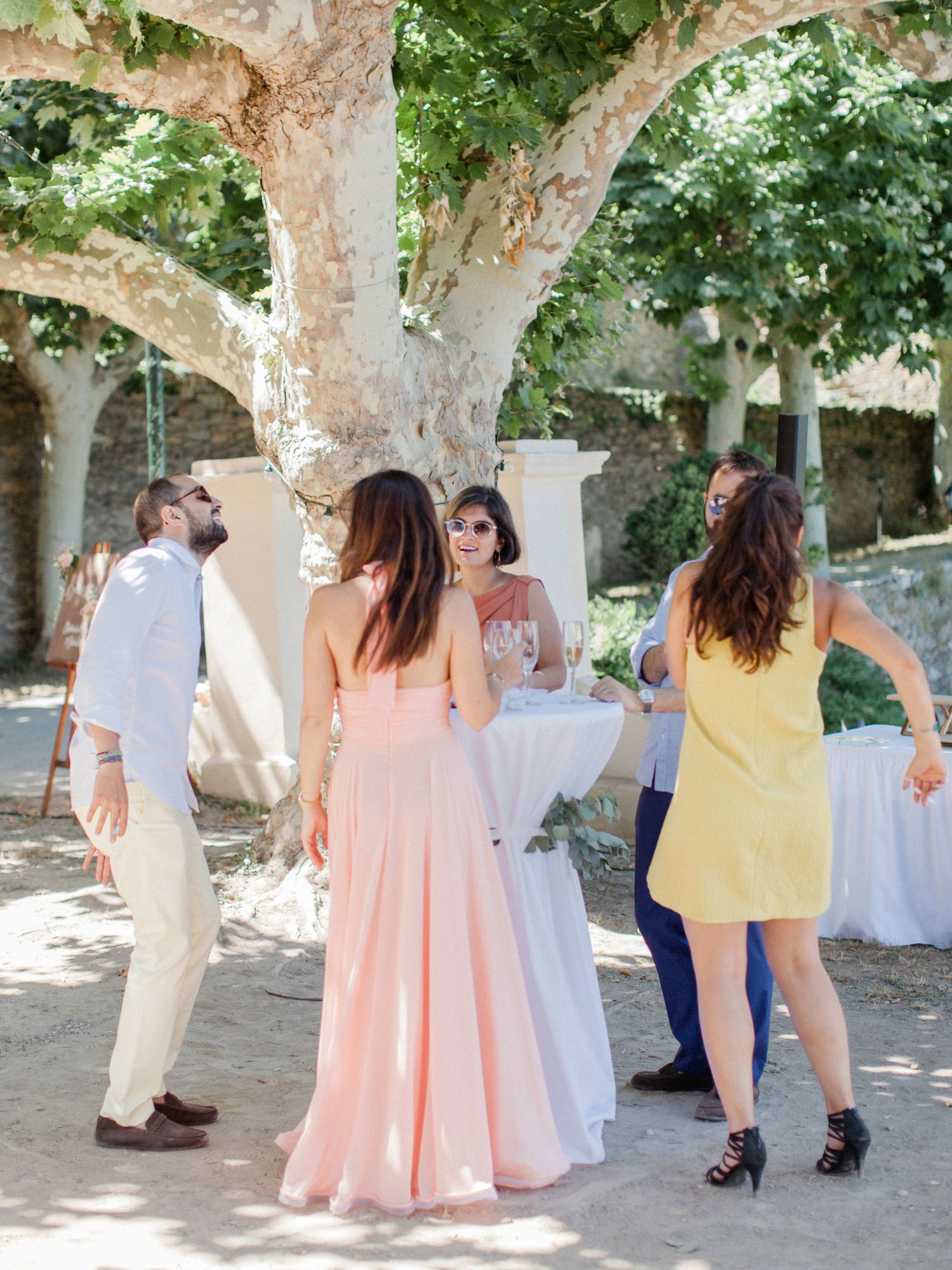 Guests in pastel dresses mingle outdoors under a large tree at an elegant summer wedding reception.