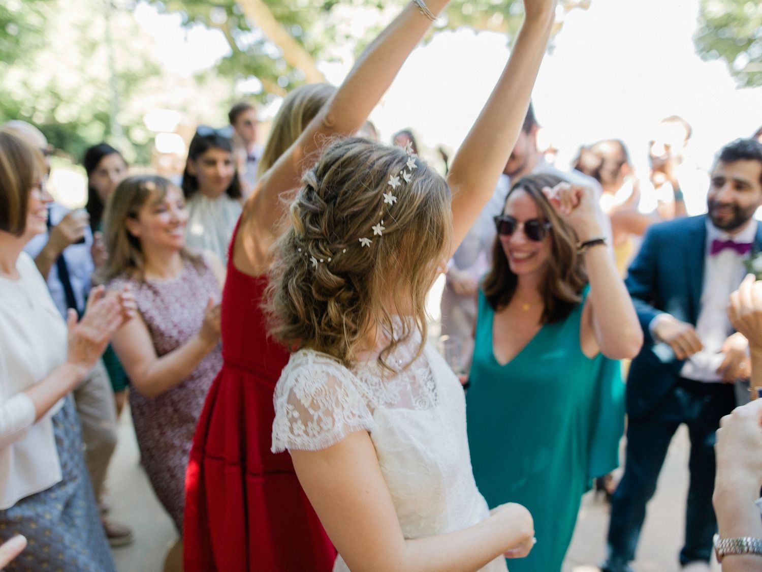 Bride in white lace dress dancing joyfully with wedding guests outdoors, arms raised in celebration.
