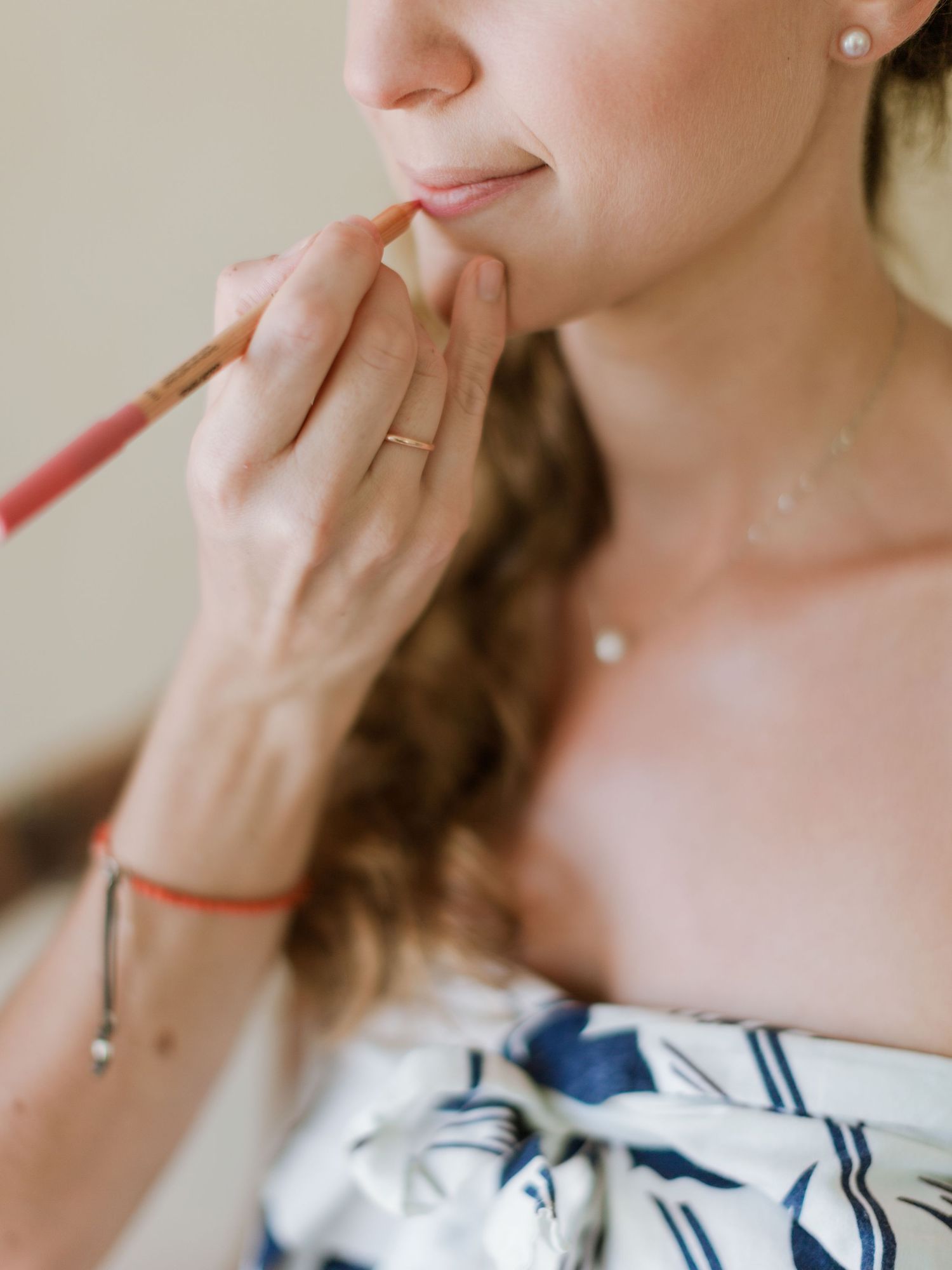 Une future mariée applique un crayon à lèvres à l'aide d'un pinceau de maquillage ; elle porte des boucles d'oreilles en perles