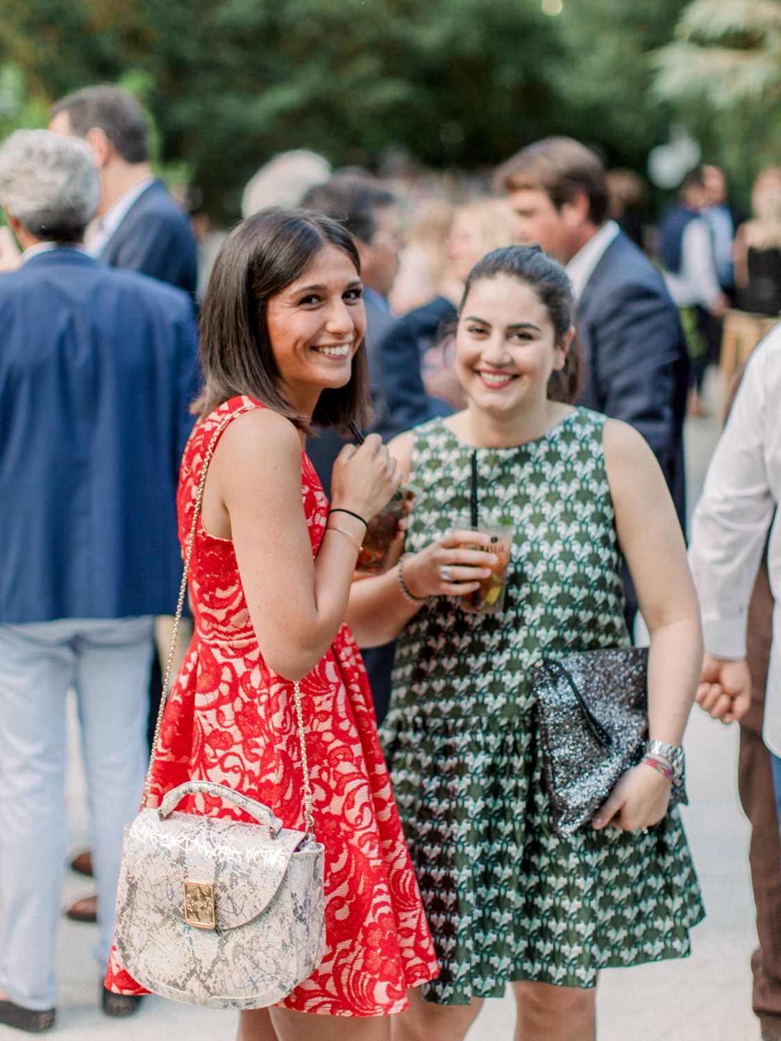 Deux femmes en robes colorées sourient lors d'une garden-party, l'une en dentelle rouge, l'autre en robe verte à motifs.