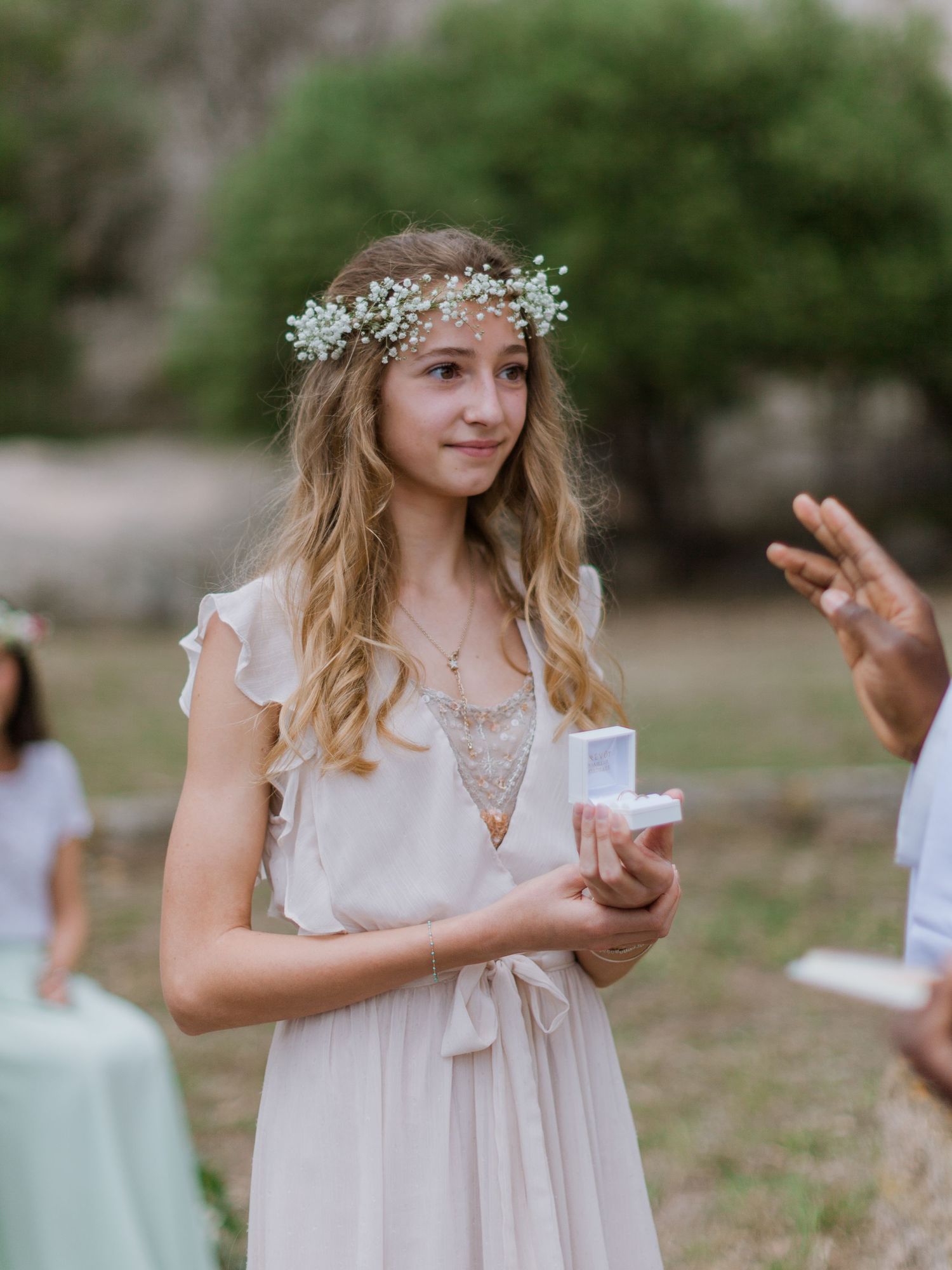 Une jeune fille en robe blanche et couronne de fleurs tient une petite boîte lors d'une cérémonie de mariage en plein air à Bonifacio