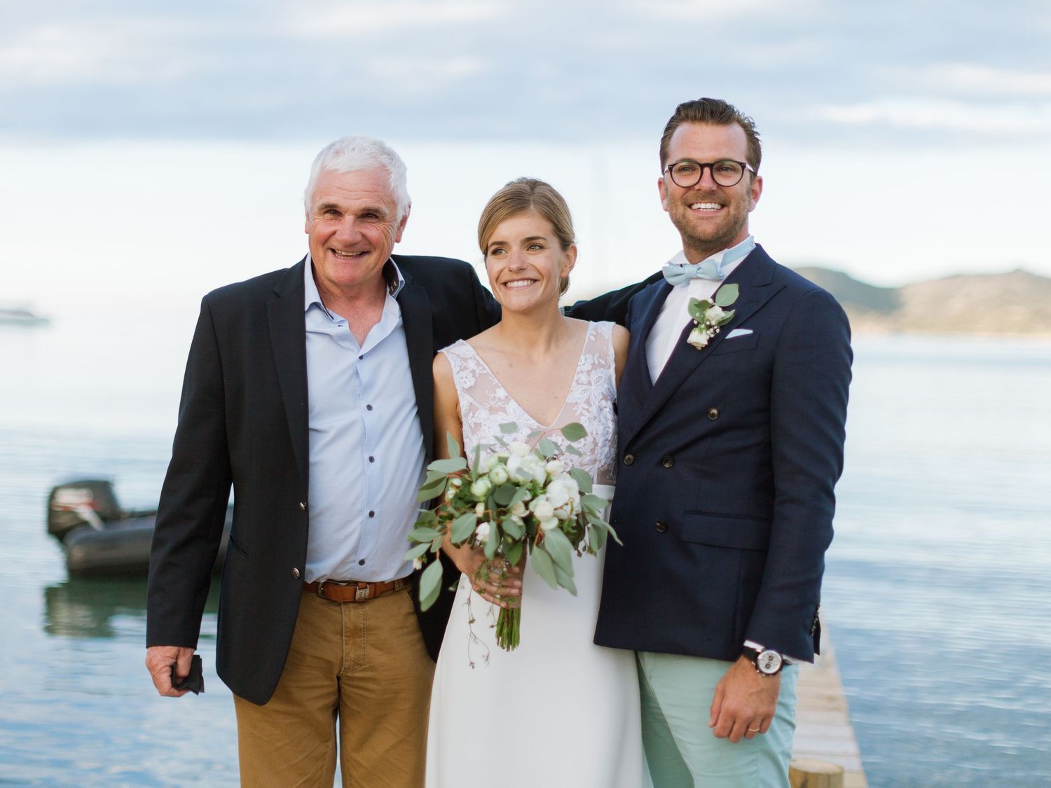 La mariée en robe blanche, tenant son bouquet, pose avec deux hommes en costume au bord de l'eau lors d'un mariage en plein air.
