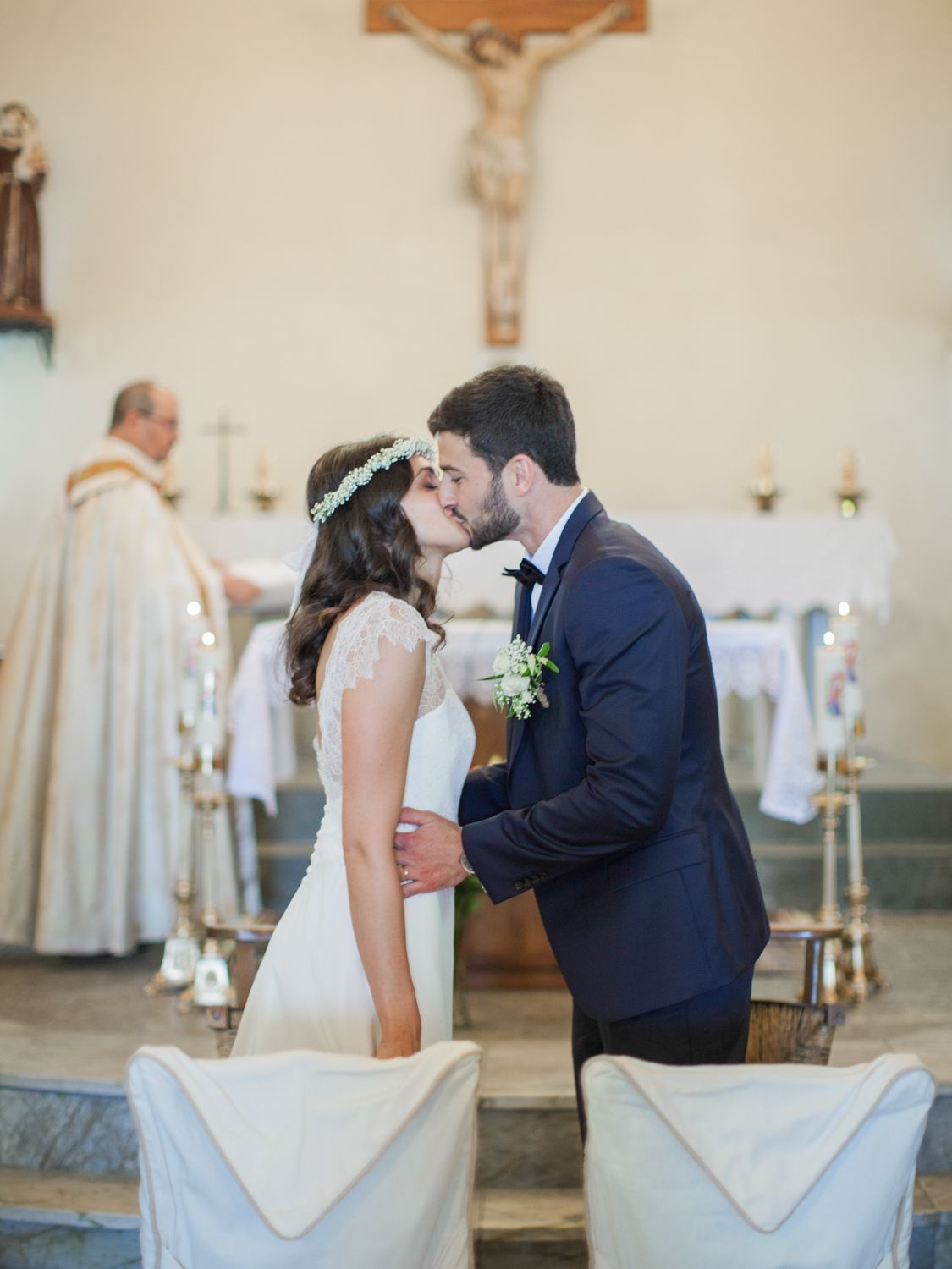 Bride and groom share first kiss at church altar during Catholic wedding ceremony with priest in background.