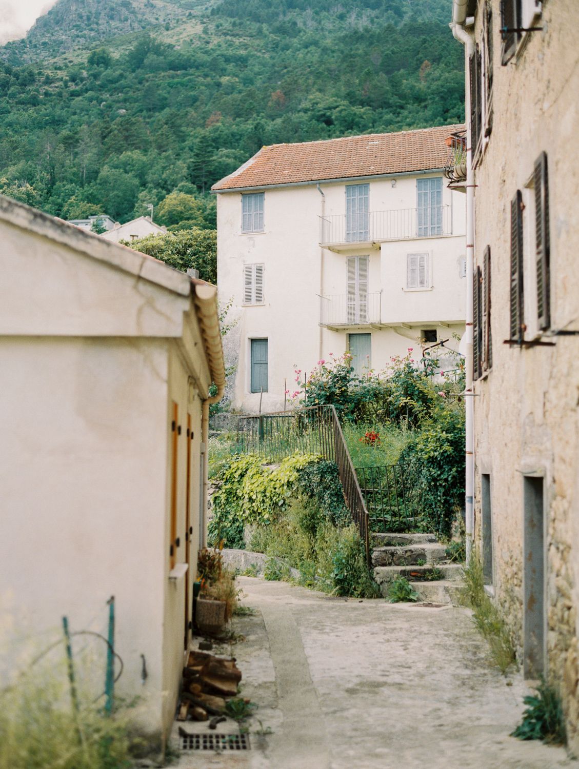 Narrow cobblestone alley between old stone buildings leading to a lush garden and white shuttered house in a French village.