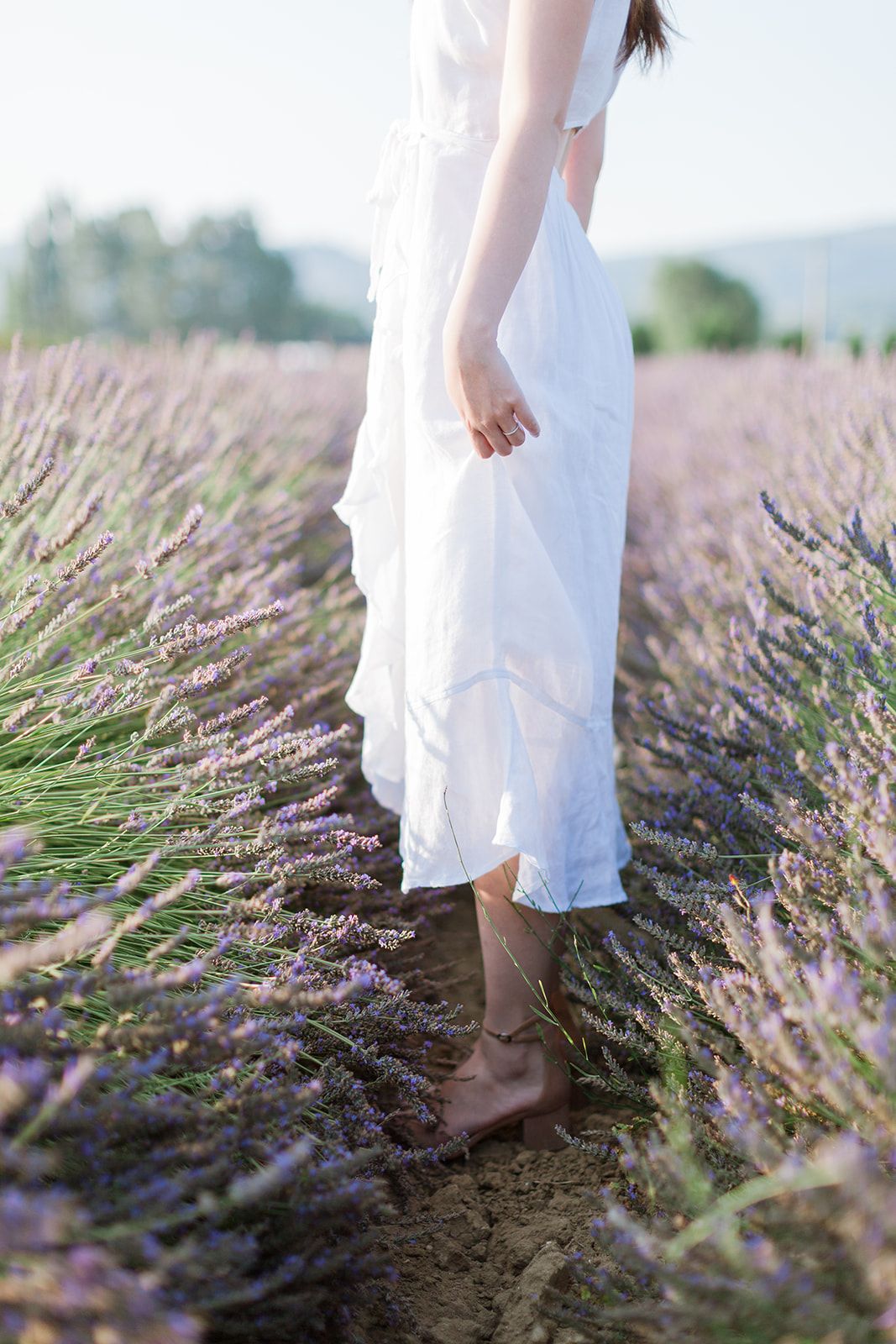 Une femme en robe blanche marche pieds nus à travers des rangées de champs de lavande violette en Provence.