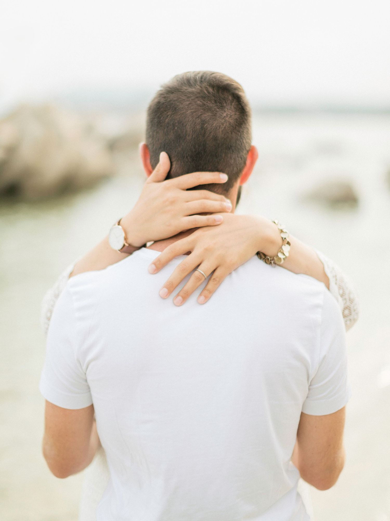 Une femme portant une bague de fiançailles enlace un homme par derrière sur une plage rocheuse. Palombaggia Porto-Vecchio.