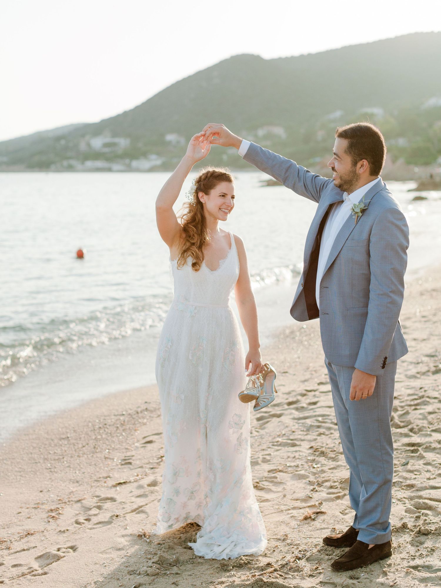 Une mariée souriante en robe de dentelle blanche est emportée par le marié en costume bleu clair sur une plage pittoresque au soleil