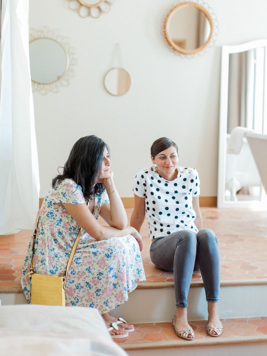 Two women sitting on steps indoors, one in a floral dress, the other in a polka dot top, engaged in conversation.
