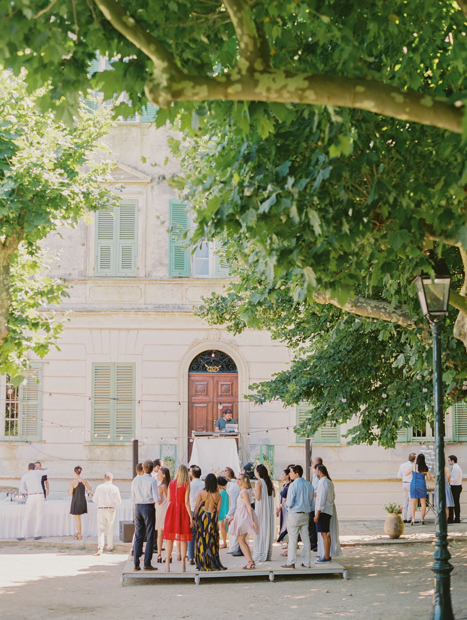 Guests gather outside a elegant French villa with green shutters and a red door, surrounded by lush trees on a sunny day.