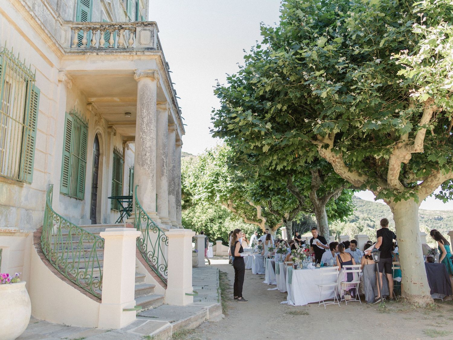 Elegant outdoor wedding reception beside a historic stone villa with guests gathered under lush green trees in warm sunlight.