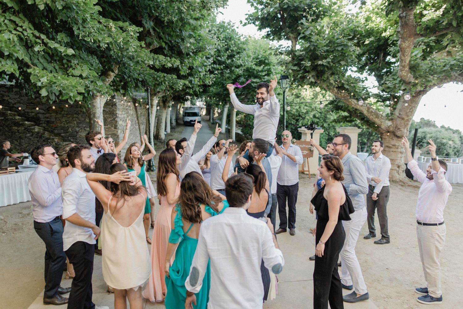 Wedding guests celebrating outdoors, with one person lifted in the air, surrounded by trees and stone walls.