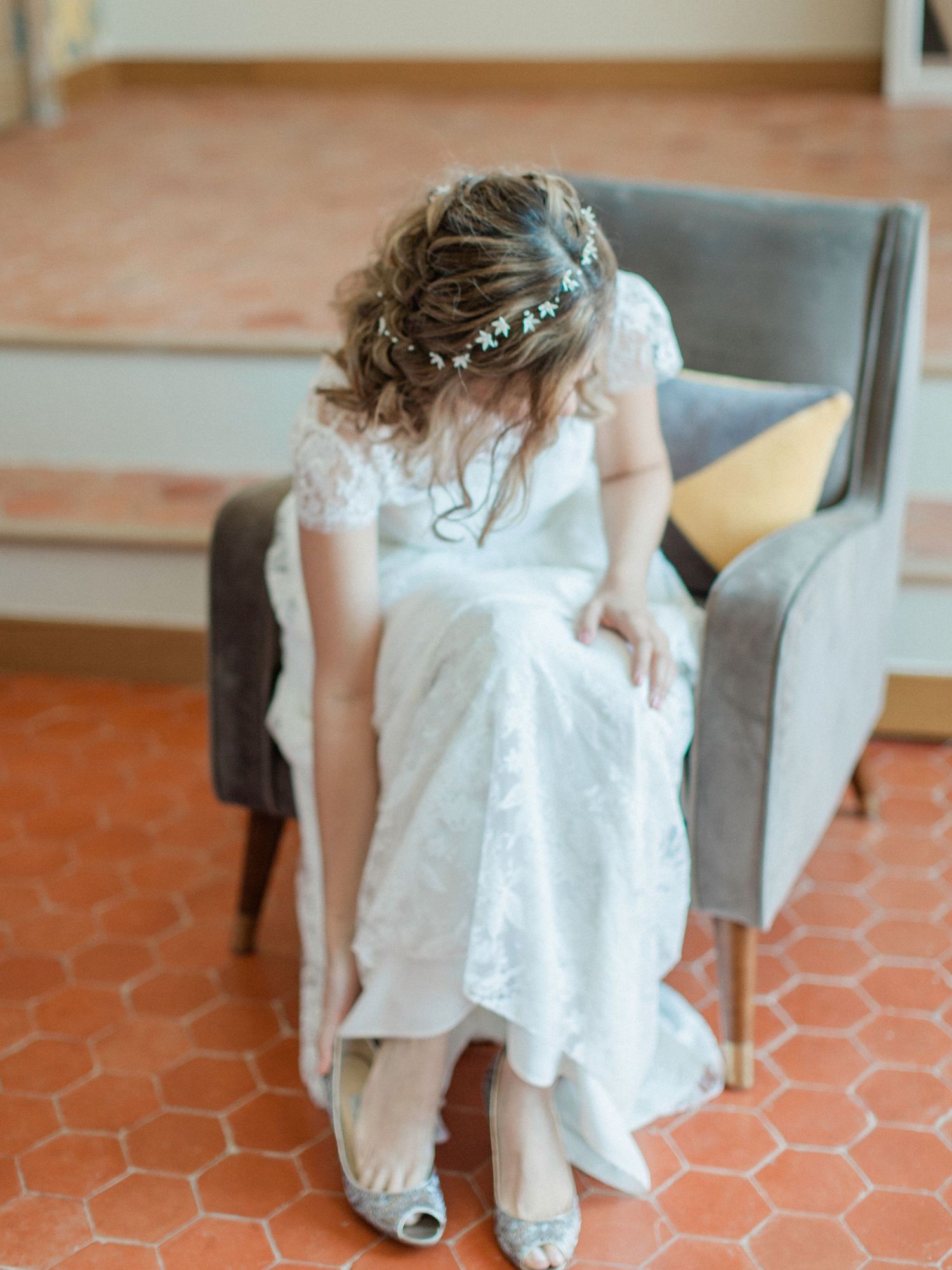Young flower girl in white lace dress with floral crown sits on gray velvet chair adjusting her silver heels.