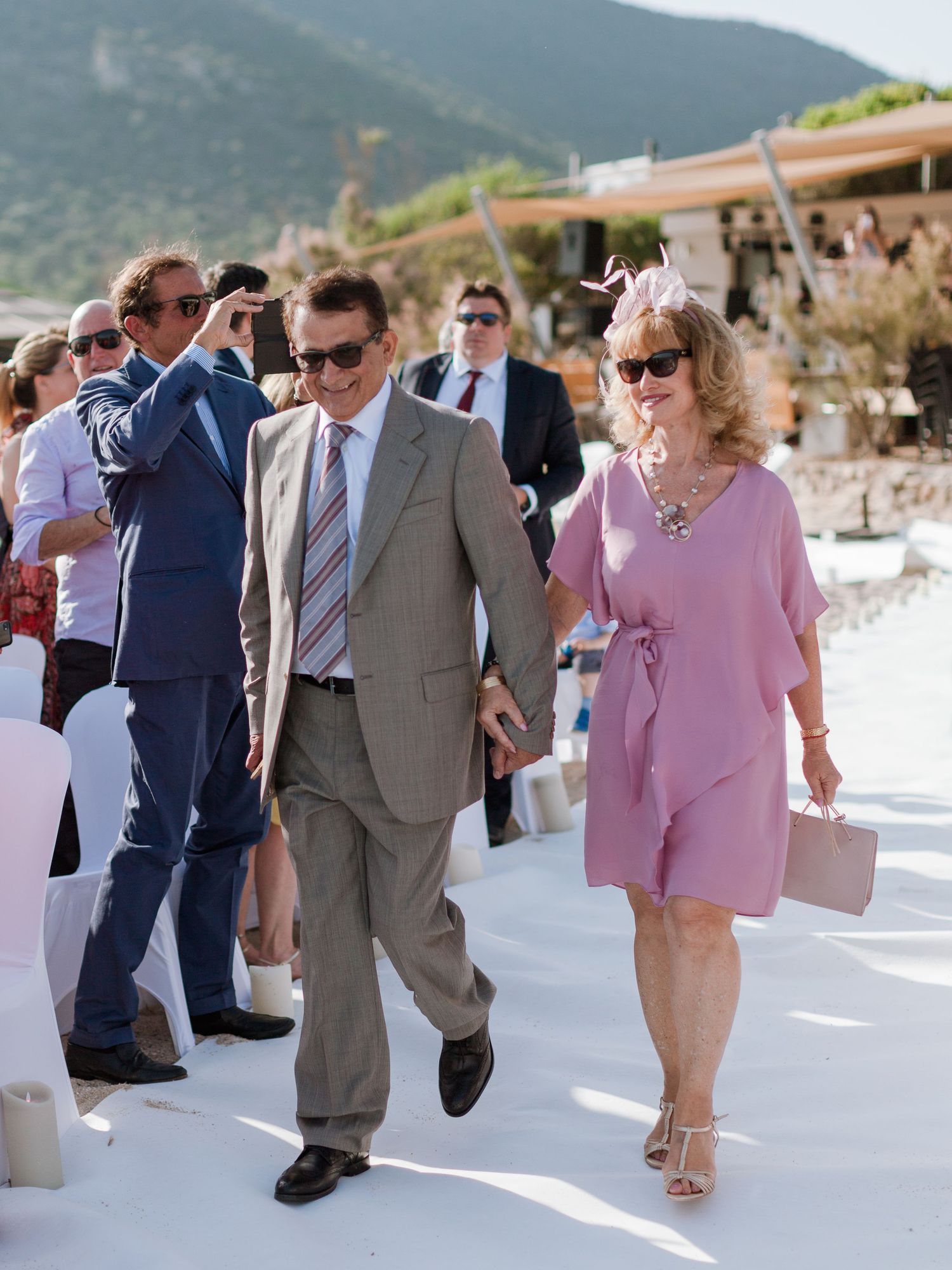 Un couple élégant marche lors d'une cérémonie de mariage en plein air ; l'homme porte un costume beige, la femme une robe rose