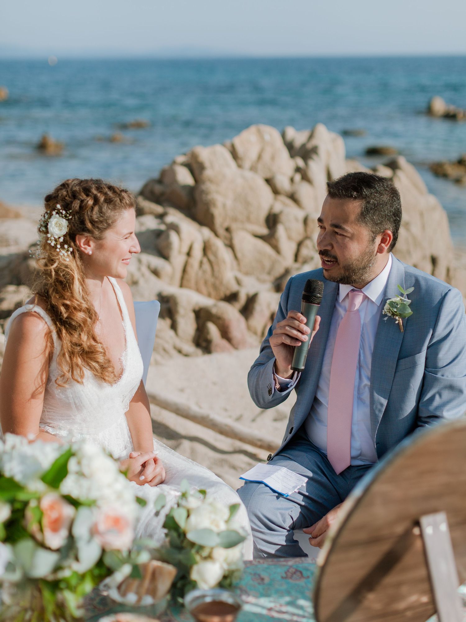 Le marié en costume bleu parle dans le microphone à la mariée en robe blanche lors d'une cérémonie de mariage romantique sur la plage.