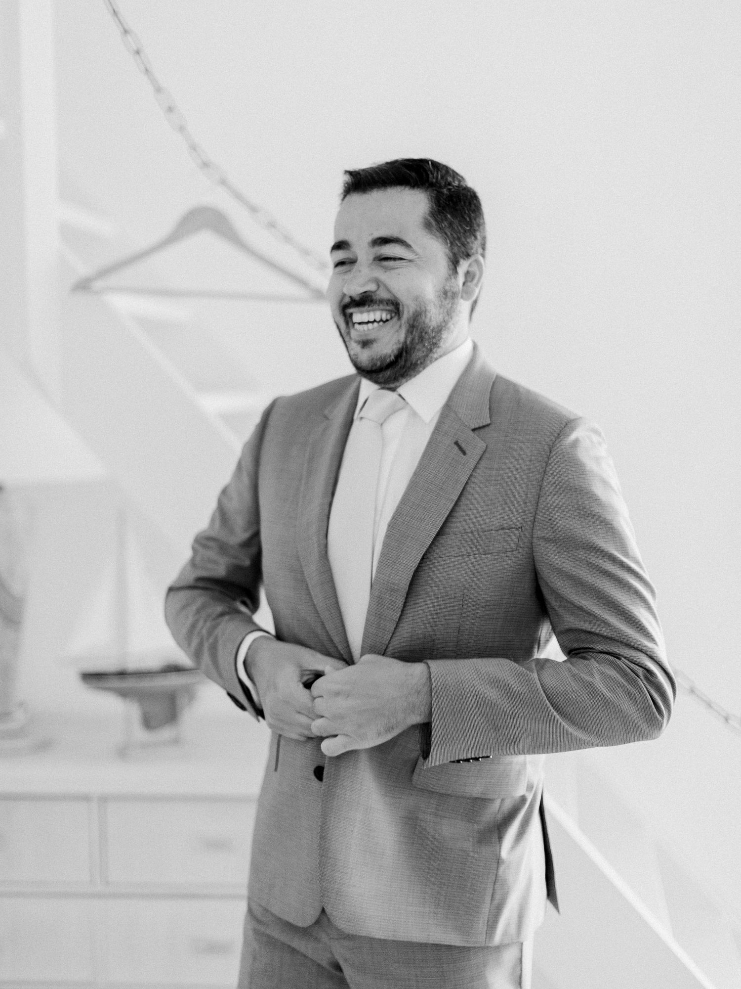 Un marié souriant, vêtu d'un costume gris, rit joyeusement alors qu'il se prépare le jour de son mariage.
