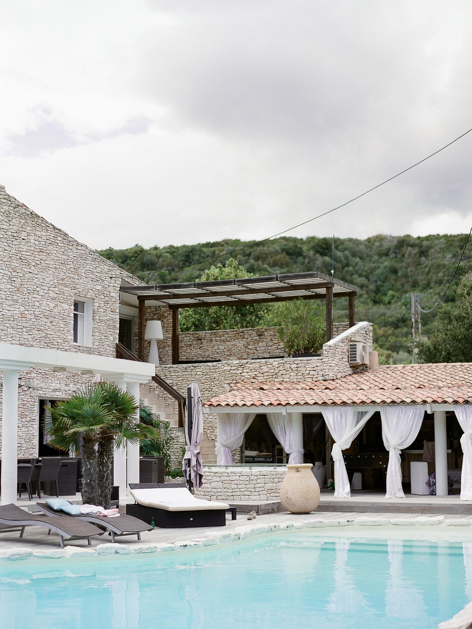 Élégante villa en pierre avec une piscine extérieure étincelante, des cabanas drapées de blanc et des collines verdoyantes en arrière-plan.