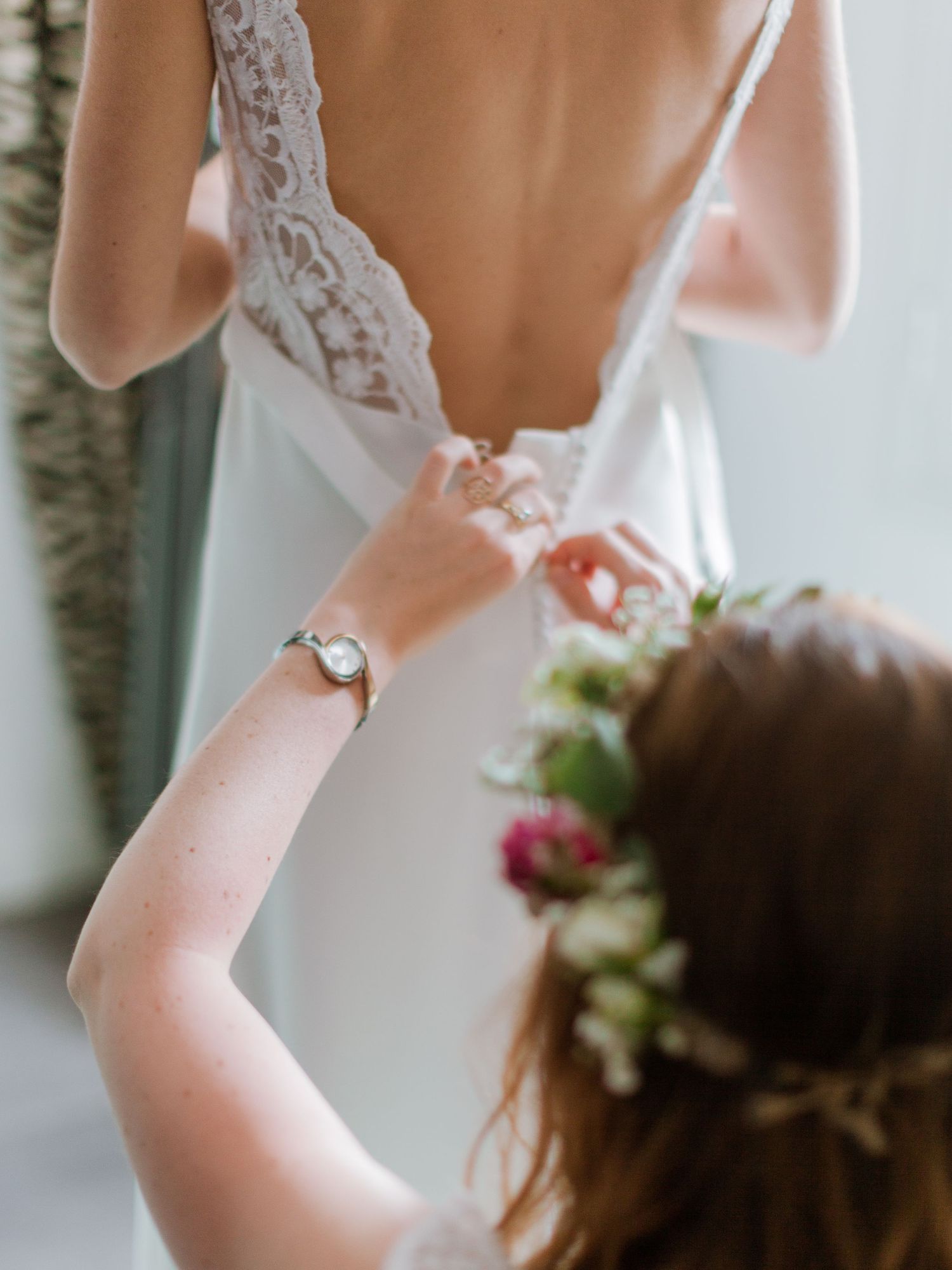 Une demoiselle d'honneur fermant le dos d'une élégante robe de mariée en dentelle, portant une couronne de fleurs et une montre en argent.