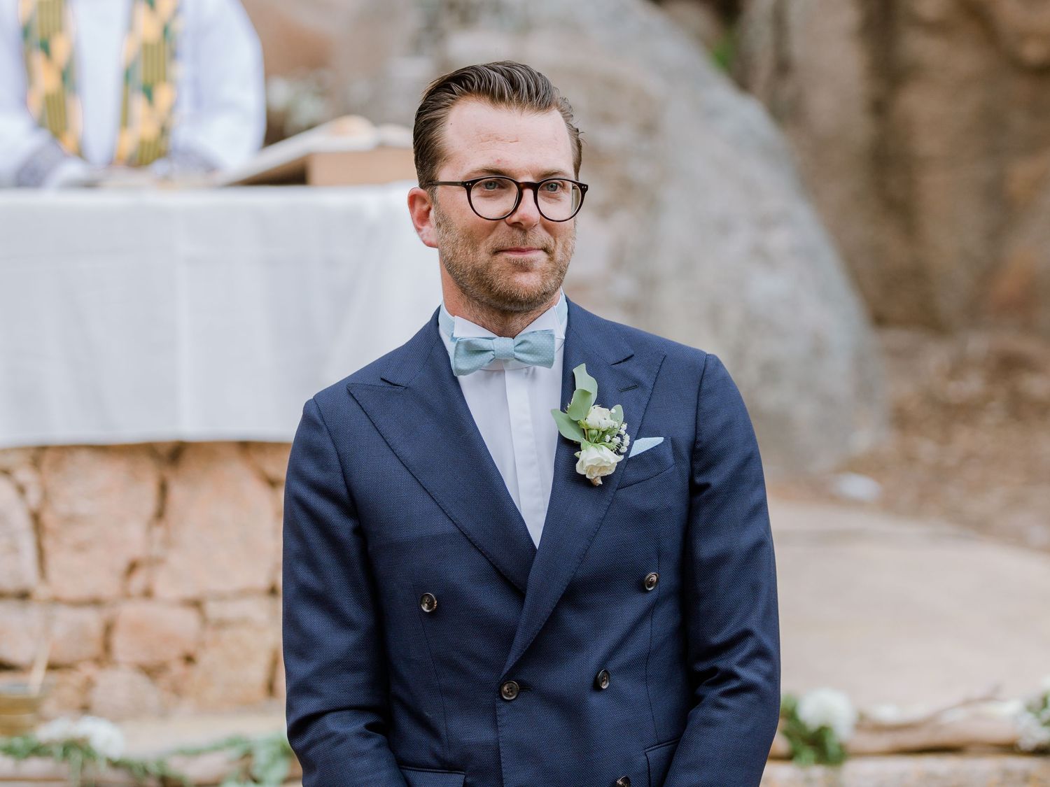 Le marié, vêtu d'un costume croisé bleu marine, avec un nœud papillon bleu clair et une boutonnière, se tient debout lors d'une cérémonie
