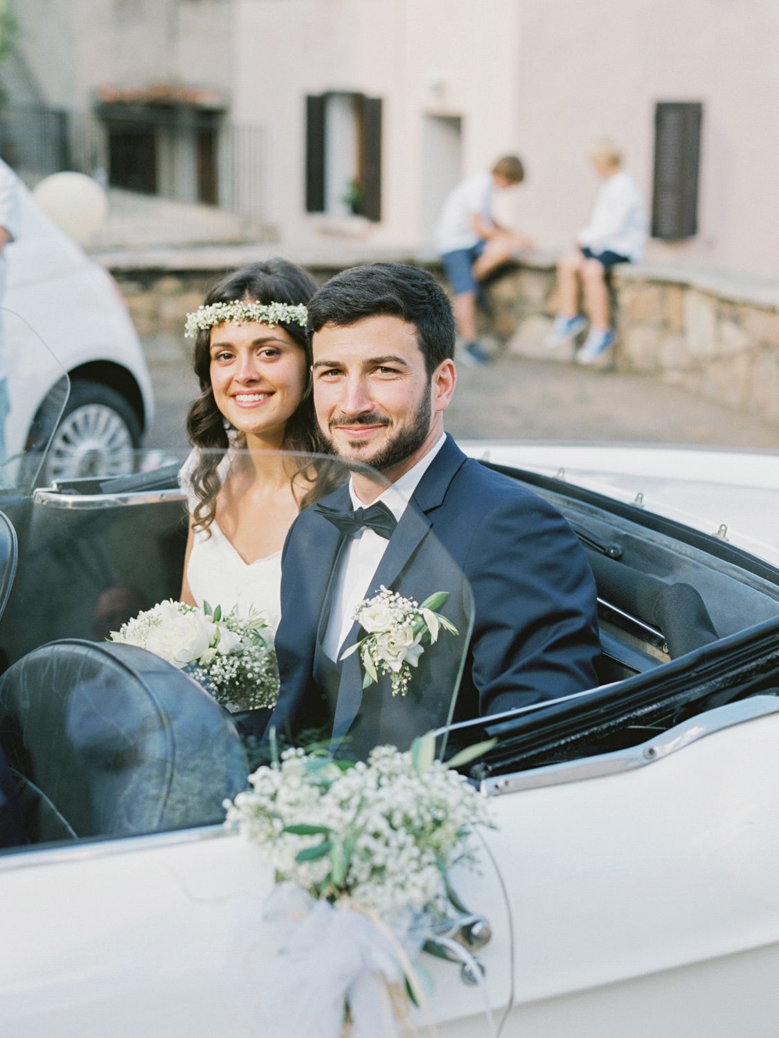 Bride and groom smiling in a white convertible car, bride wearing floral crown, groom in navy suit with boutonniere.