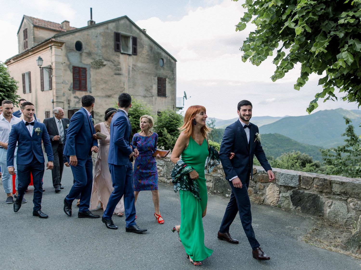 Wedding party walking along a stone path with scenic mountain views and a historic building in Tuscany, Italy.