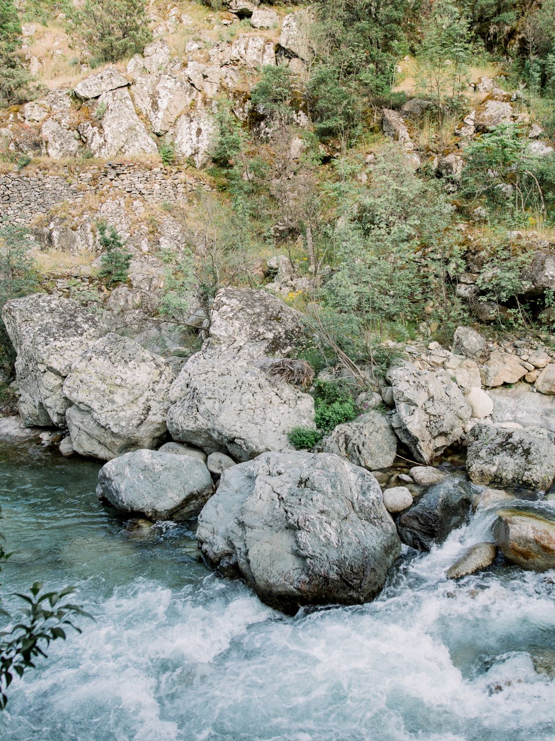 Rocky mountain river with rushing white water rapids flowing around large boulders, surrounded by lush green trees and rocky cliffs.