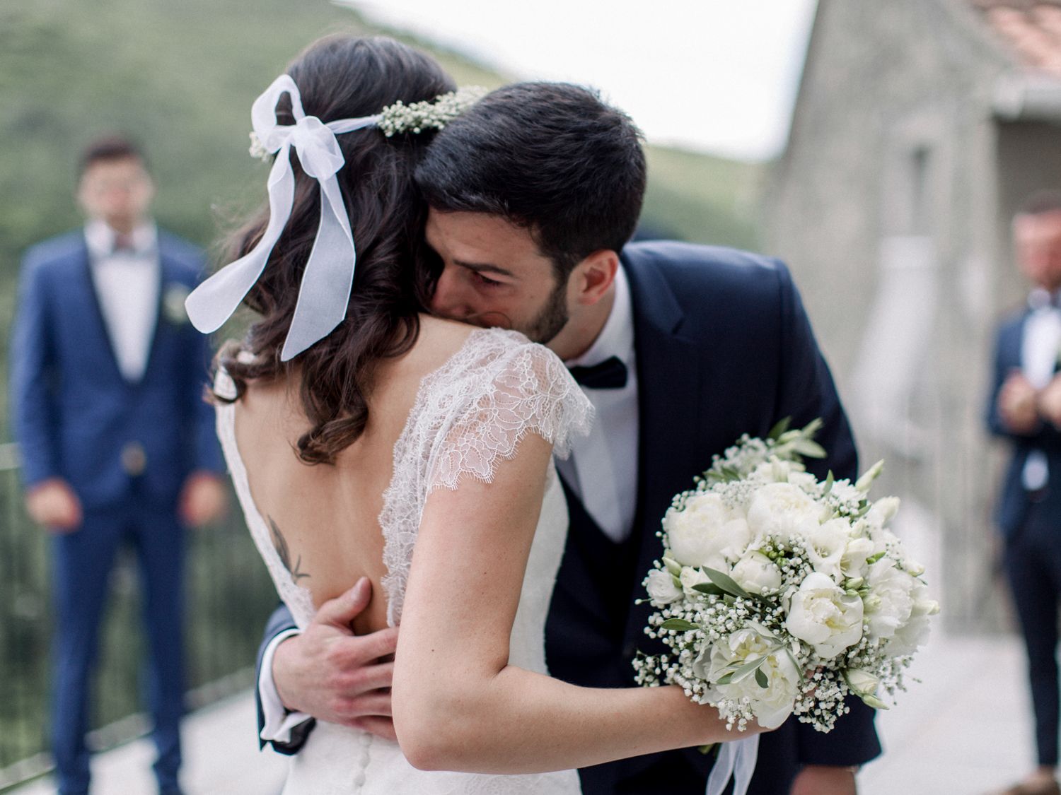 A bride and groom share a romantic embrace during their outdoor wedding ceremony, with the bride holding a white floral bouquet.