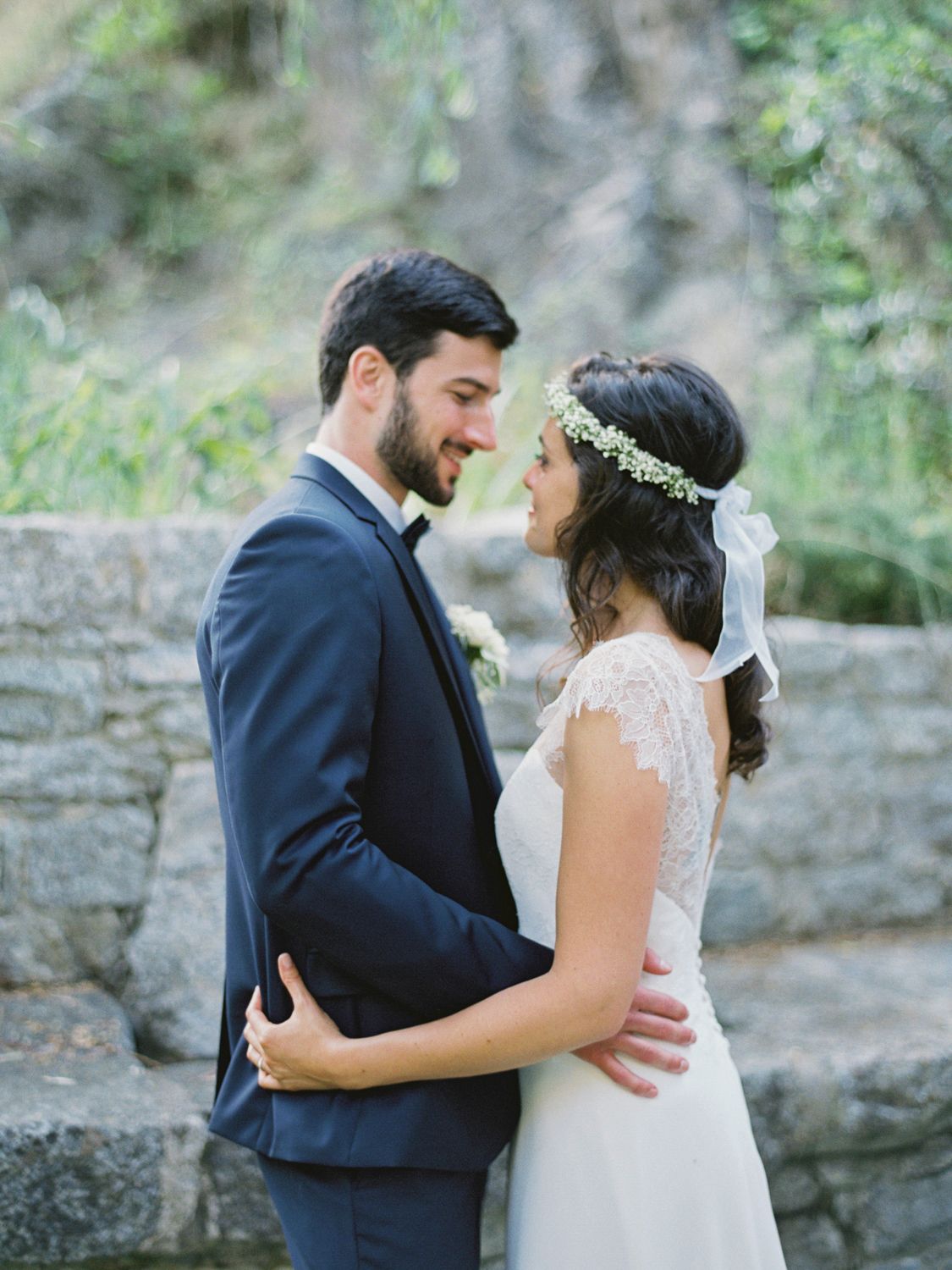 A bride and groom share a romantic moment in wedding attire, she wears a floral crown and lace dress outdoors.