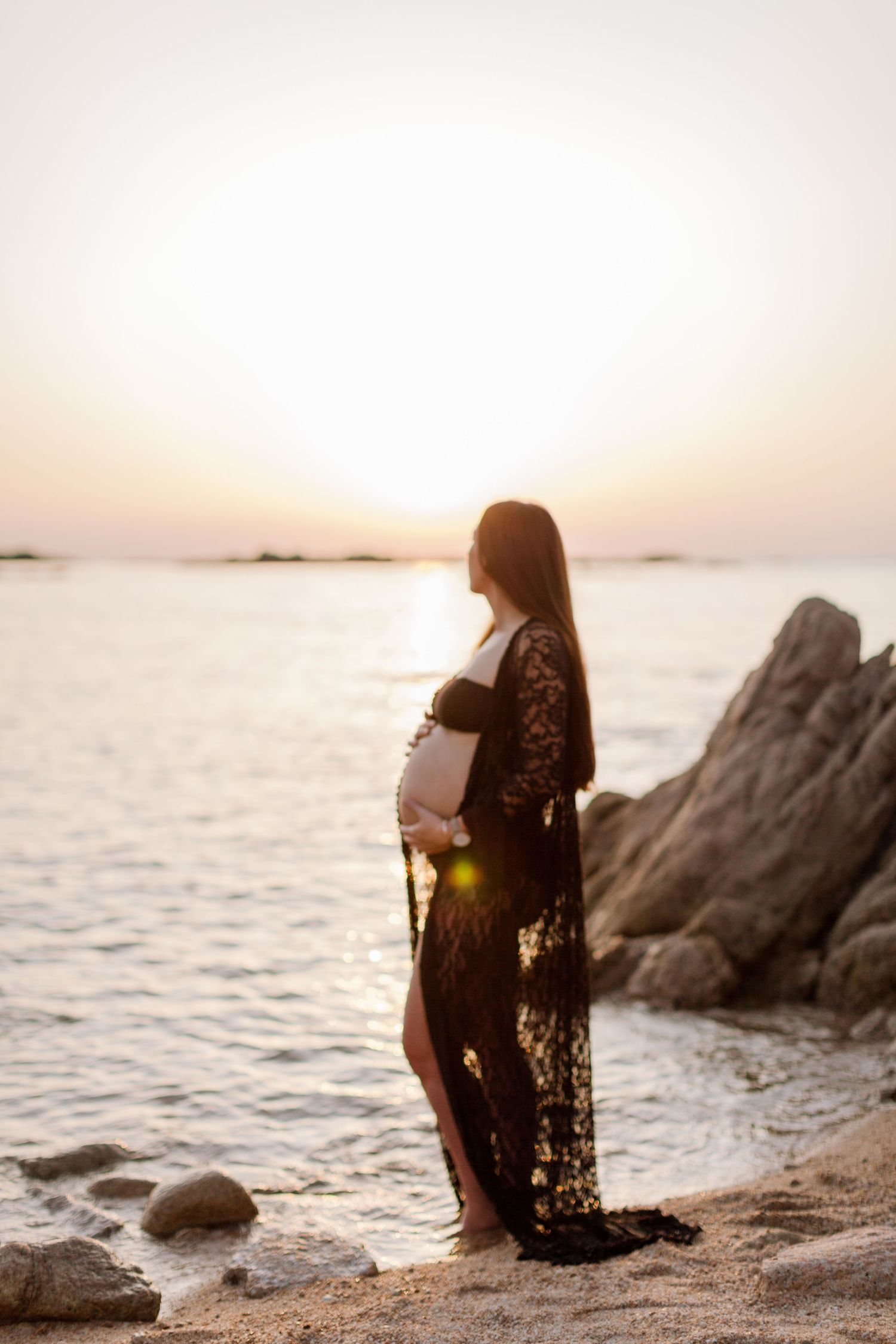 Femme enceinte en robe de dentelle noire, berçant son ventre sur une plage rocheuse au coucher du soleil doré en bord de mer à Bonifacio.