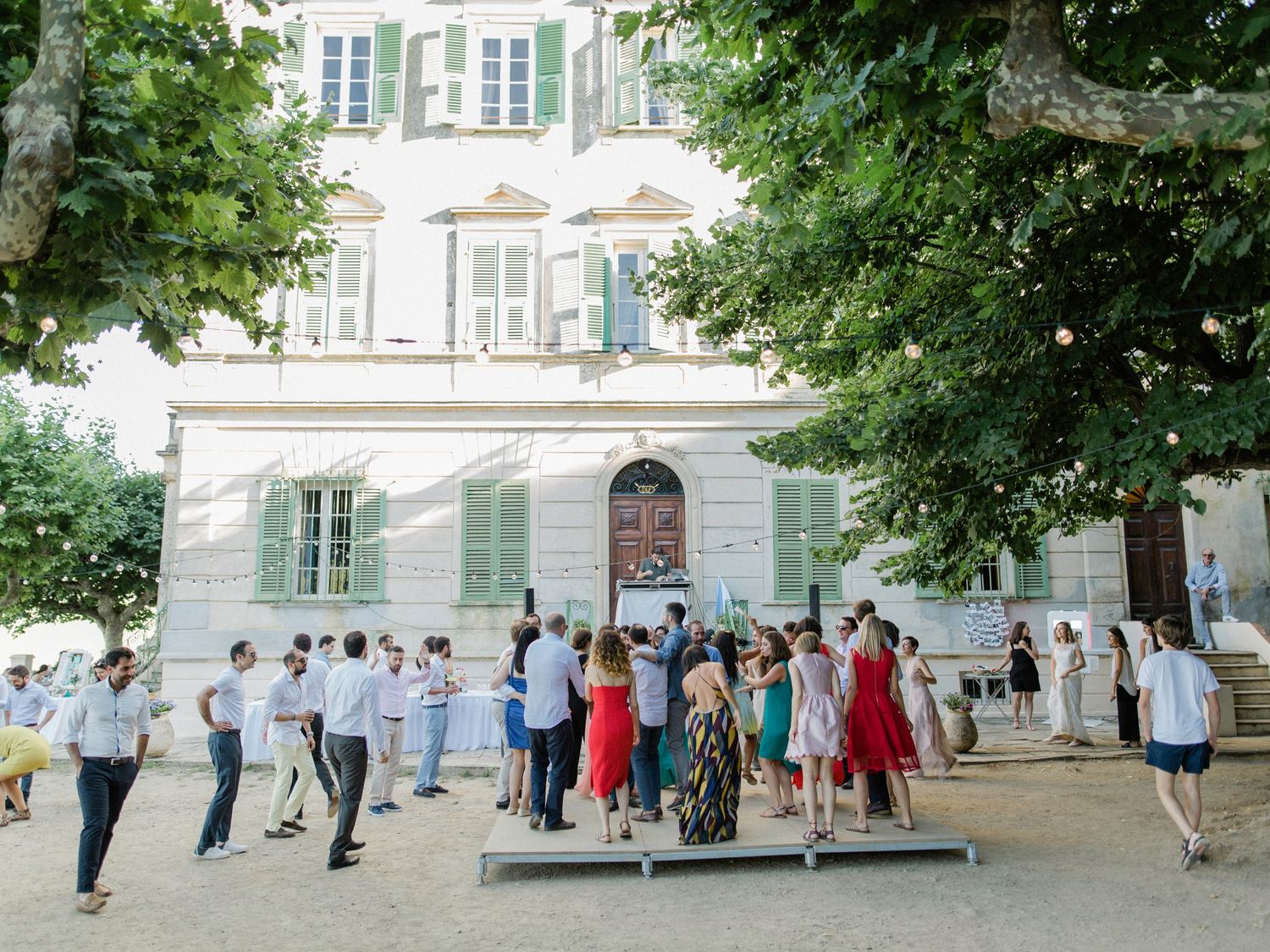 Guests mingle outside a elegant French villa with green shutters during an outdoor summer gathering.