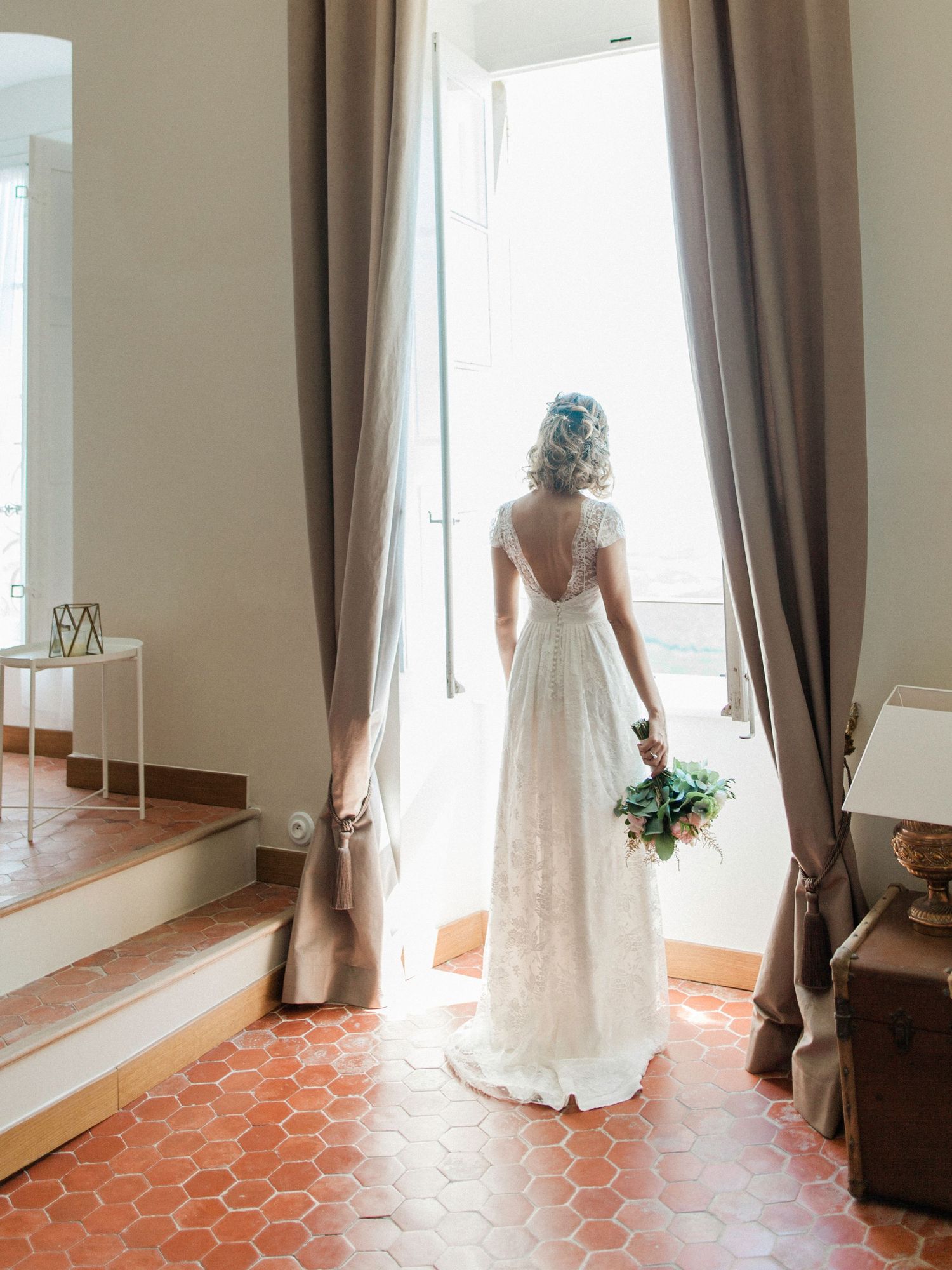 Bride in lace wedding gown holding bouquet stands at open French doors with terracotta tile floor.