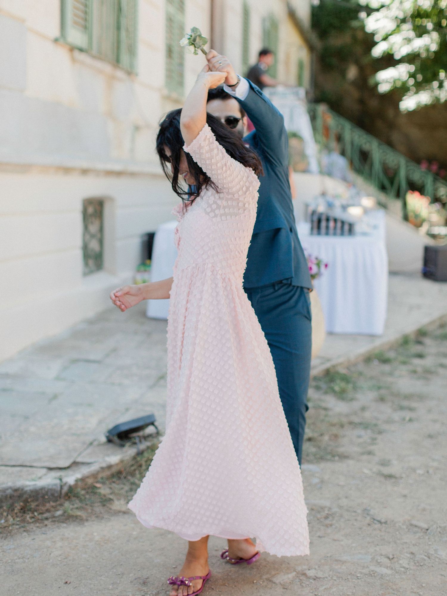 Couple dancing outdoors at a wedding, woman in white polka dot dress twirling, man in teal suit, elegant venue backdrop.