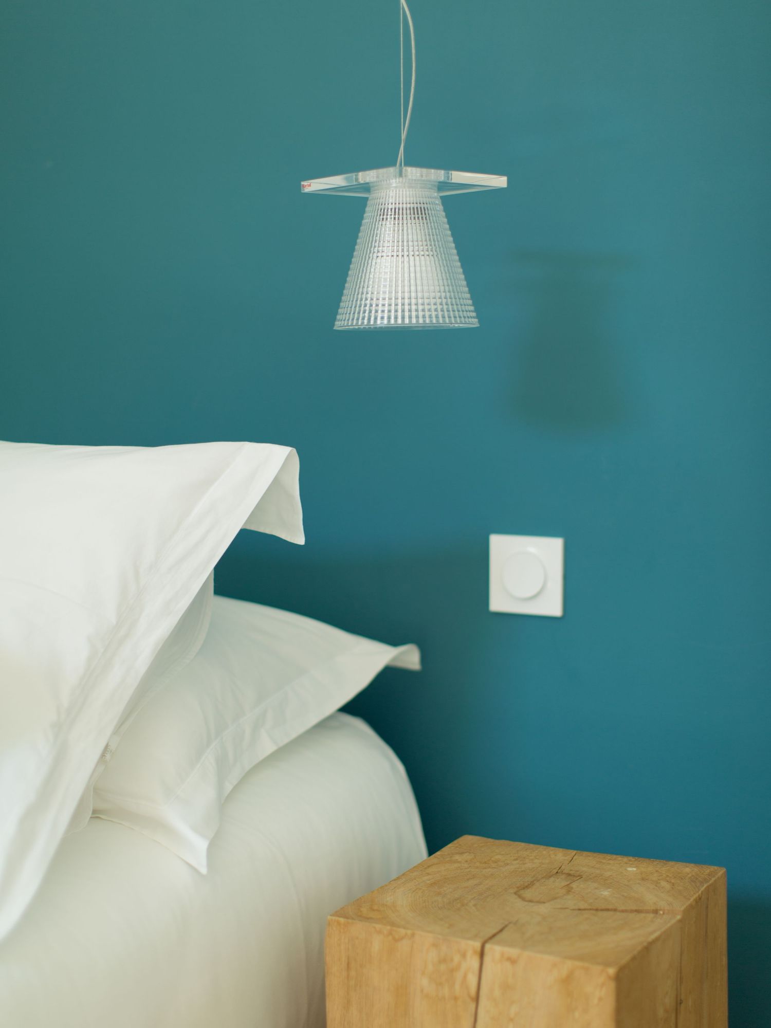 Mur bleu sarcelle dans la chambre, avec une suspension transparente à nervures au-dessus d'oreillers blancs et d'une table de chevet en bois