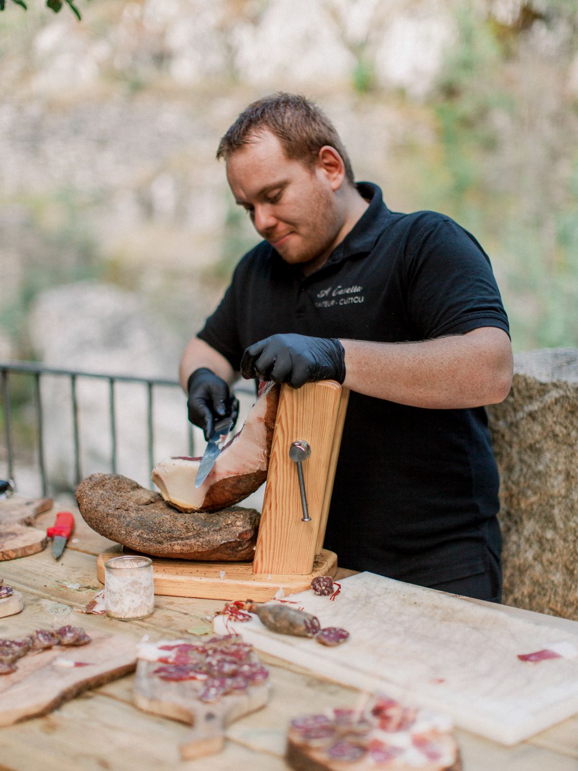 Un chef ganté de noir découpe de la charcuterie sur une planche en bois à un stand de charcuterie en plein air.